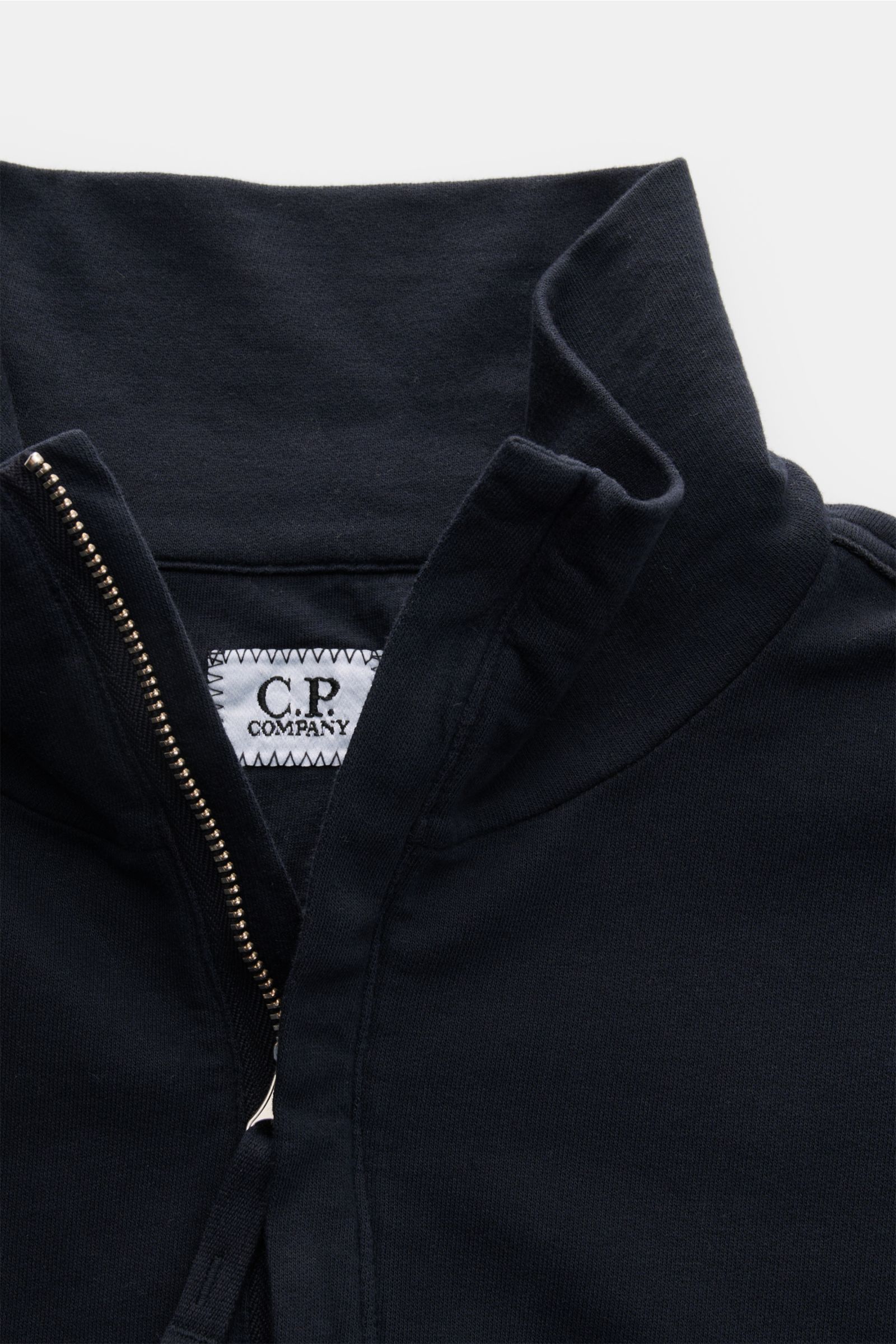 Close-up image of the C.P. Company Sweat-Troyer navy from a top front perspective, showing the high collar with a partially unzipped zipper and the C.P. Company label inside the collar. Schon auf den ersten Blick spürt man die sportive DNA: Das innovative