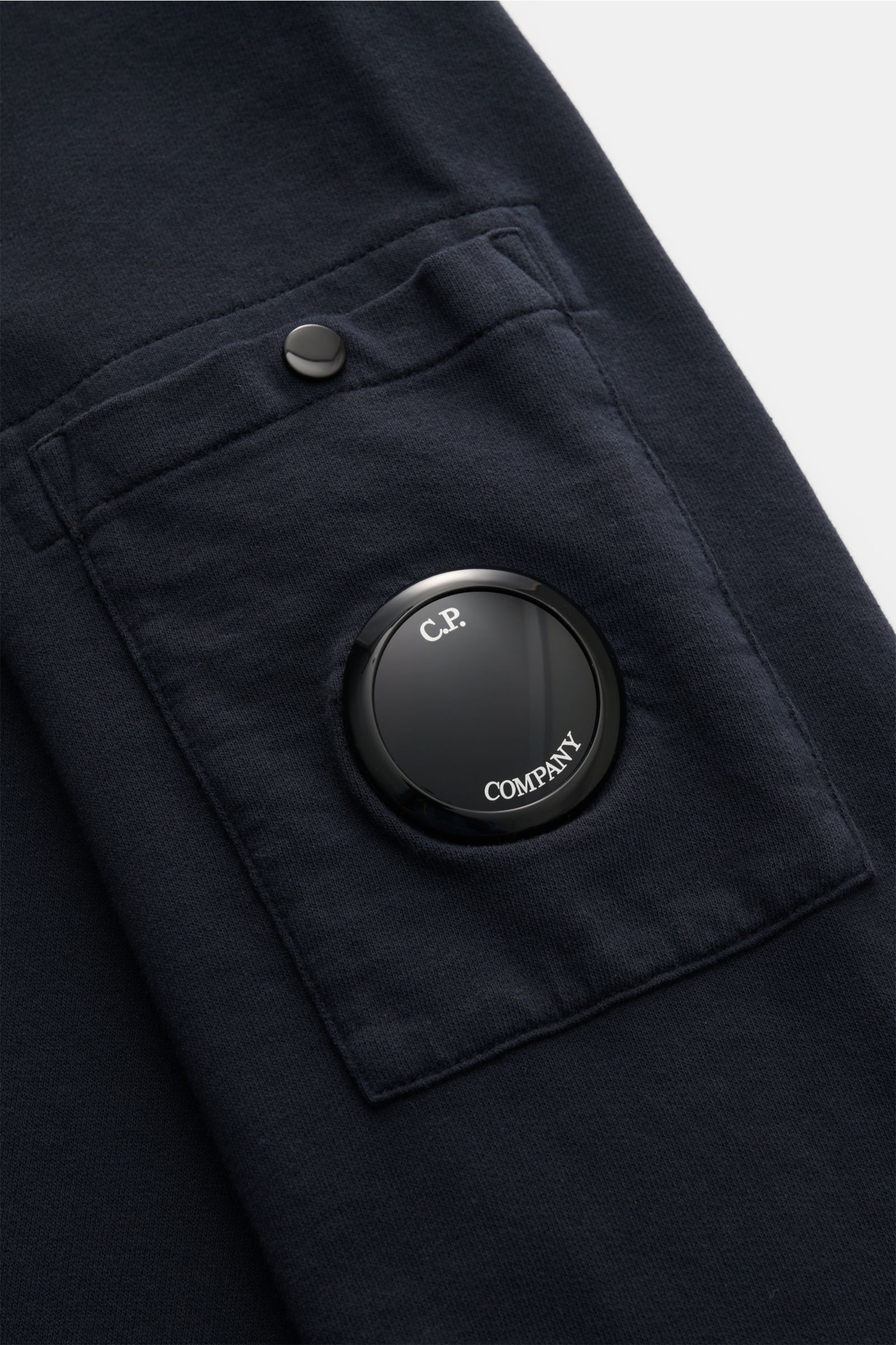 Close-up side view of the C.P. Company Sweat-Troyer navy sleeve pocket featuring a black lens detail and snap button on soft, lightweight cotton fabric. Schon auf den ersten Blick spürt man die sportive DNA: Das innovative Casualwear-Label C.P. COMPANY pr