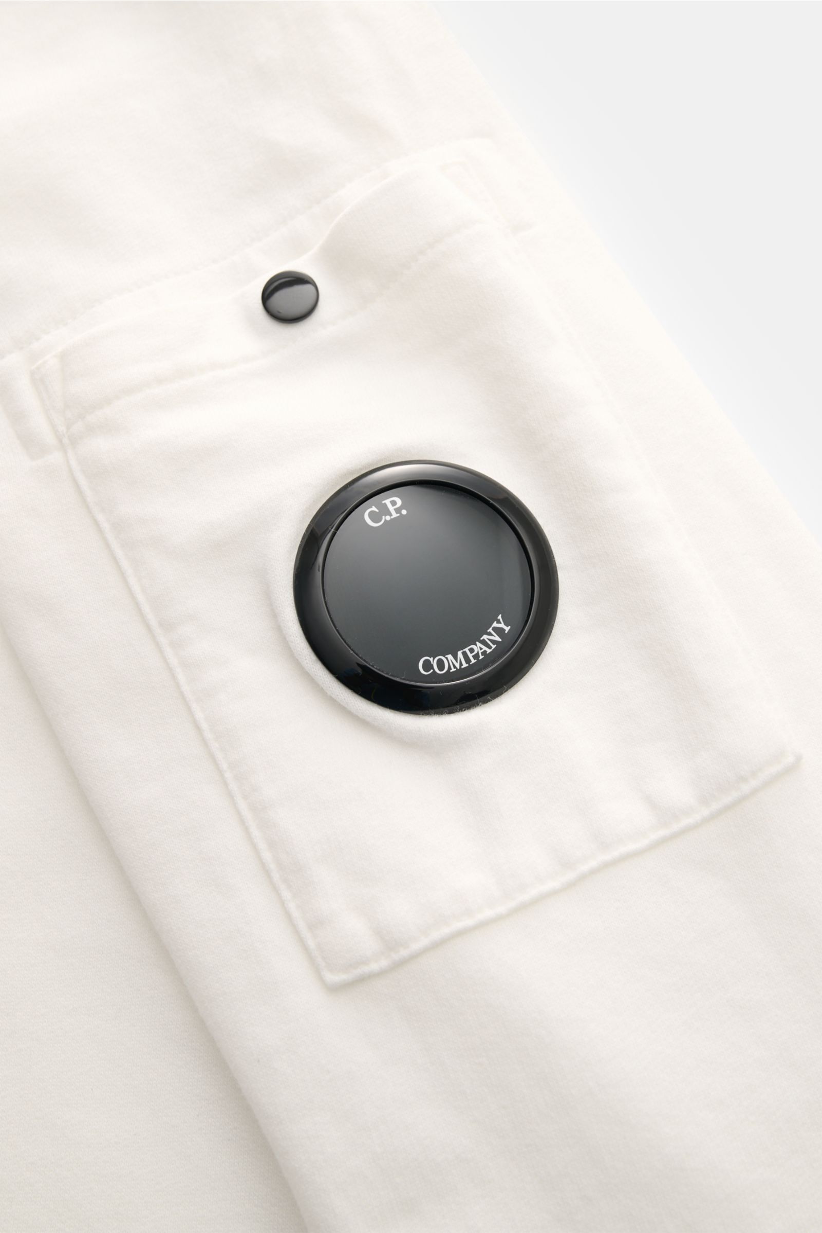 Close-up front-side view of C.P. Company Sweat-Troyer offwhite pocket with black lens detail on offwhite cotton fabric.

Incorporated content summary: Schon auf den ersten Blick spürt man die sportive DNA: Das innovative Casualwear-Label C.P. COMPANY präs