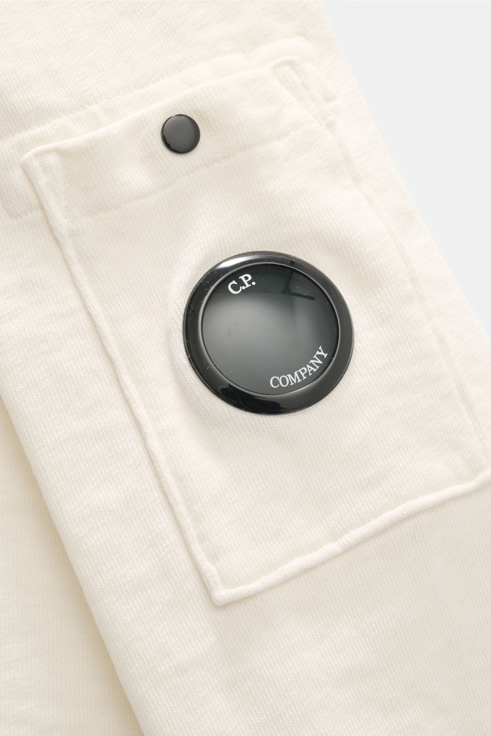Nahaufnahme des C.P. Company Rundhals-Sweatshirts creme, Perspektive auf die Oberarmtasche mit Druckknopf und Lens-Detail.