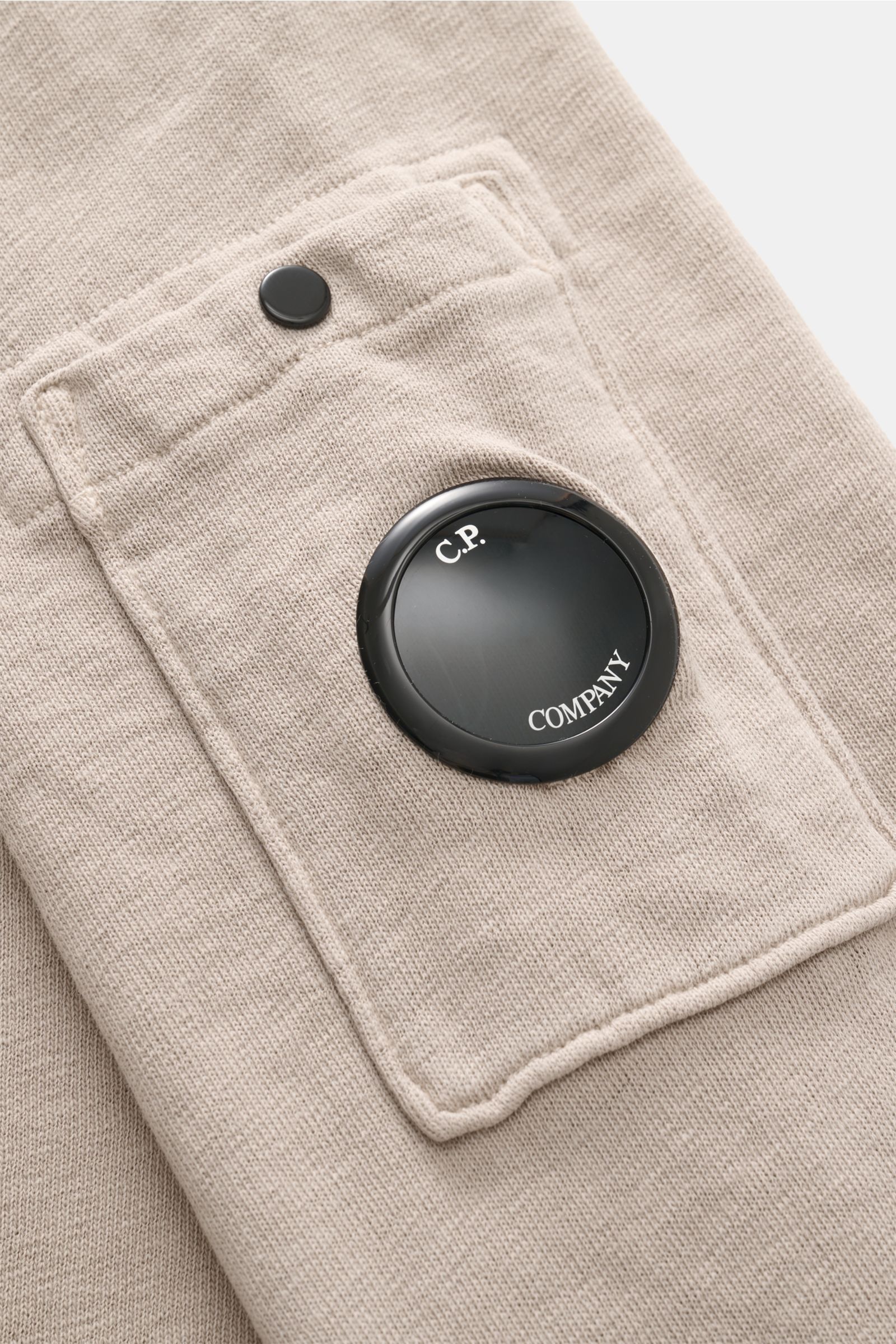 Close-up view of the C.P. Company Rundhals-Sweatshirt beige, showing the cotton fabric pocket with a black lens detail and snap button; 