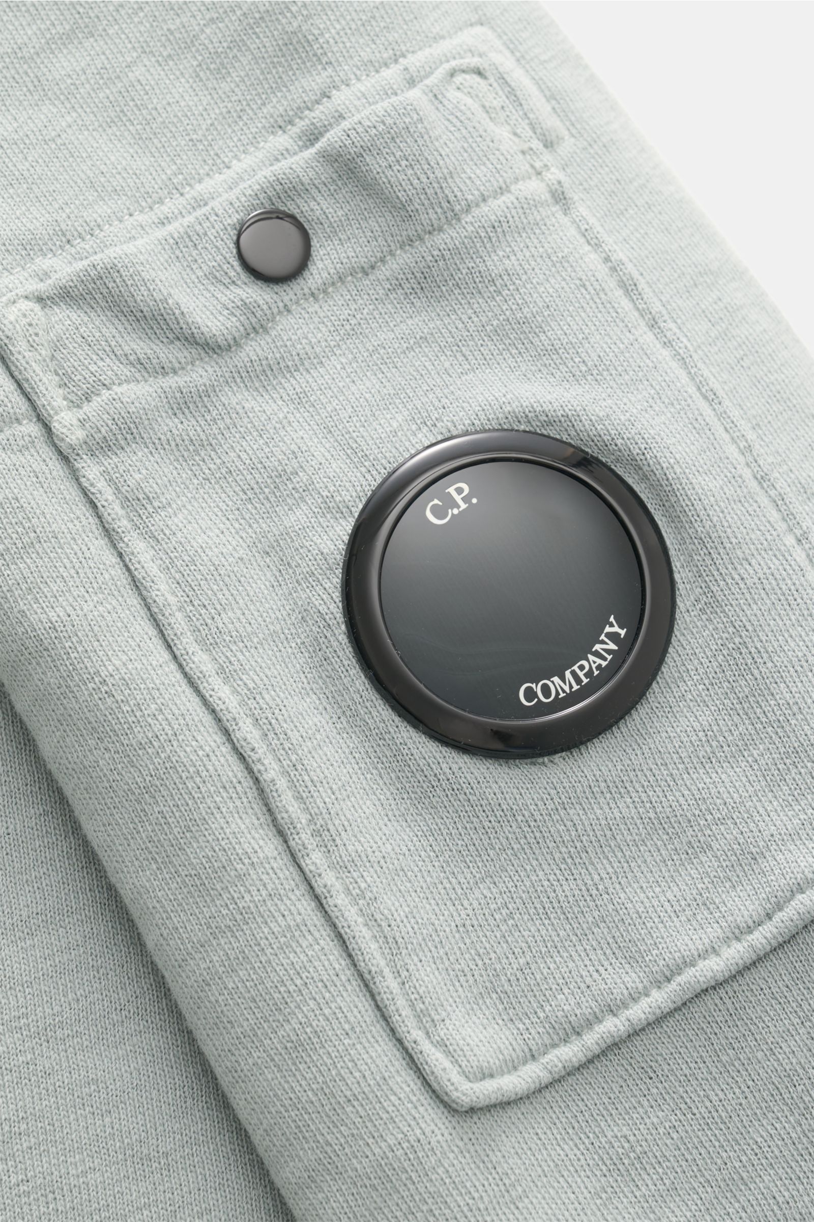 Close-up front-side view of C.P. Company Crew neck sweatshirt grey-green showing garment-dyed cotton fabric, snap button pocket, and signature lens detail. 