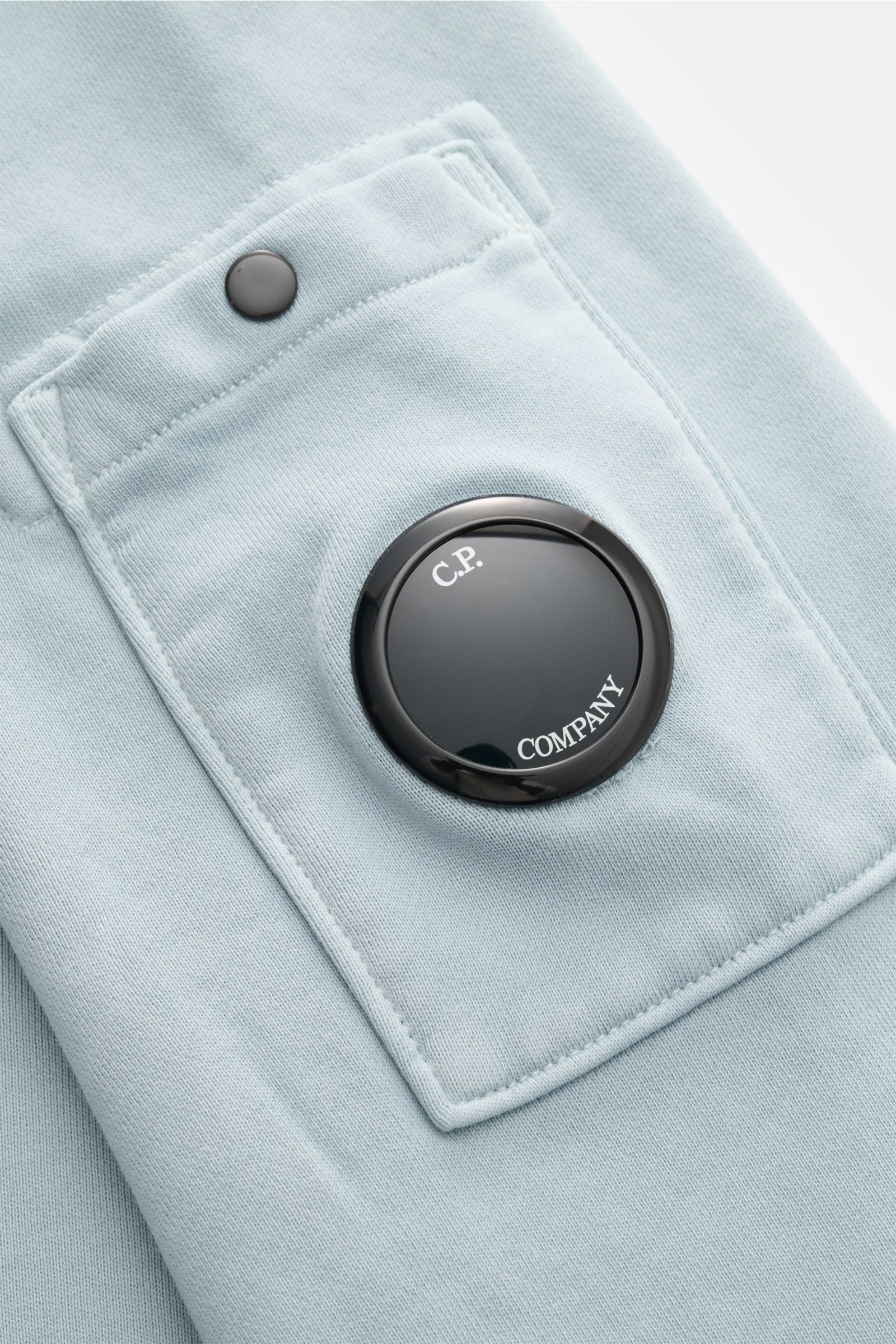 Nahaufnahme des rauchblauen C.P. Company Sweatshirts aus reiner Baumwolle, seitlich fotografiert, Fokus auf Ärmeltasche mit schwarzem Linsen-Detail.