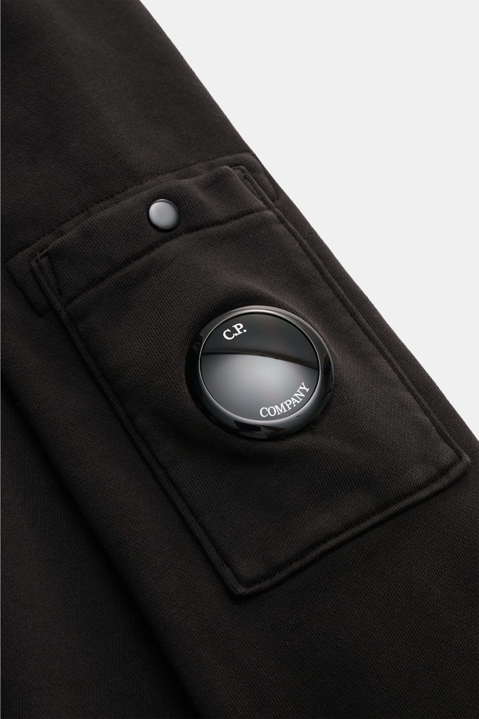Nahaufnahme des schwarzen C.P. Company Sweatshirts, Perspektive auf die Ärmeltasche mit labeltypischem Linsen-Detail aus glänzendem Material.