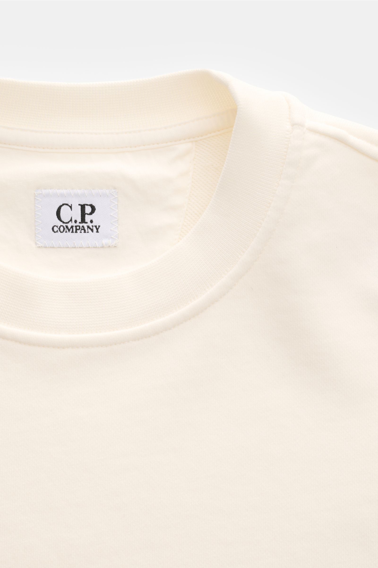 Nahaufnahme des offwhite C.P. Company Sweatshirts mit Rundhalsausschnitt und weißem Logo-Label innen, fotografiert von oben.