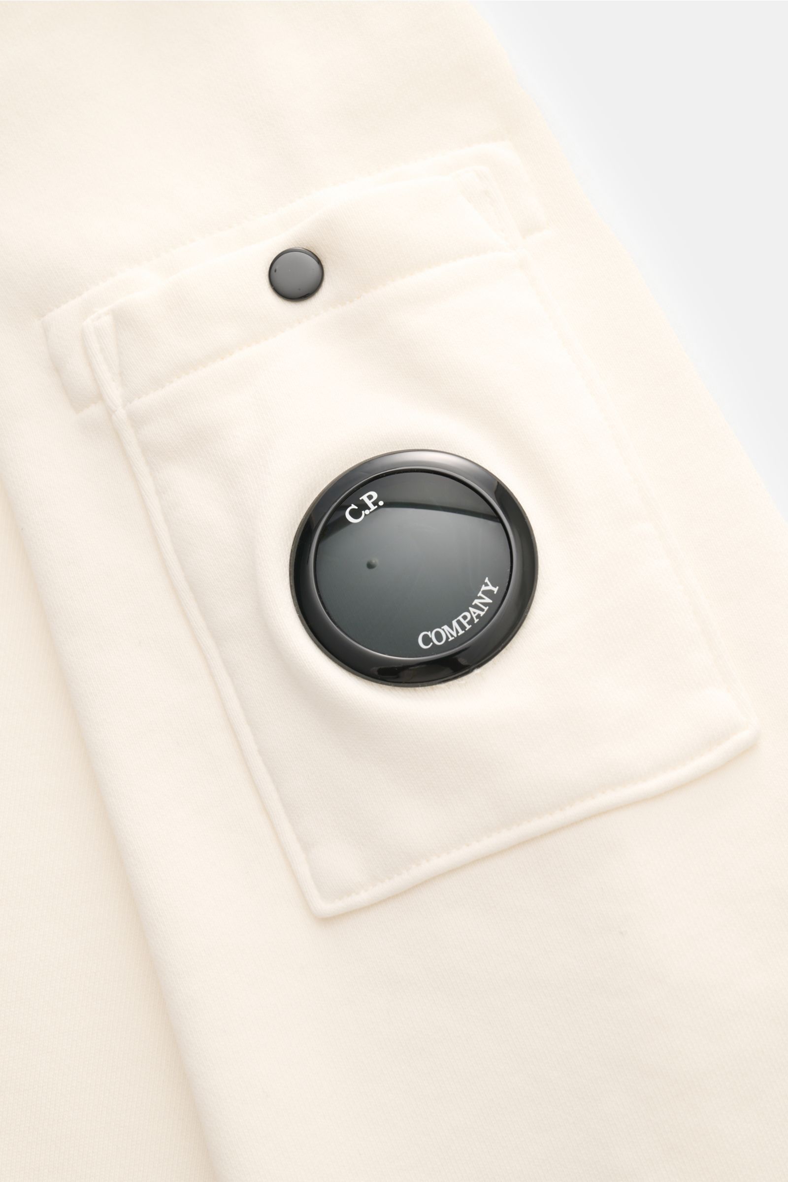 Nahaufnahme des offwhite C.P. Company Sweatshirts von schräg oben, Fokus auf die Ärmeltasche mit labeltypischem schwarzem Linsen-Detail.