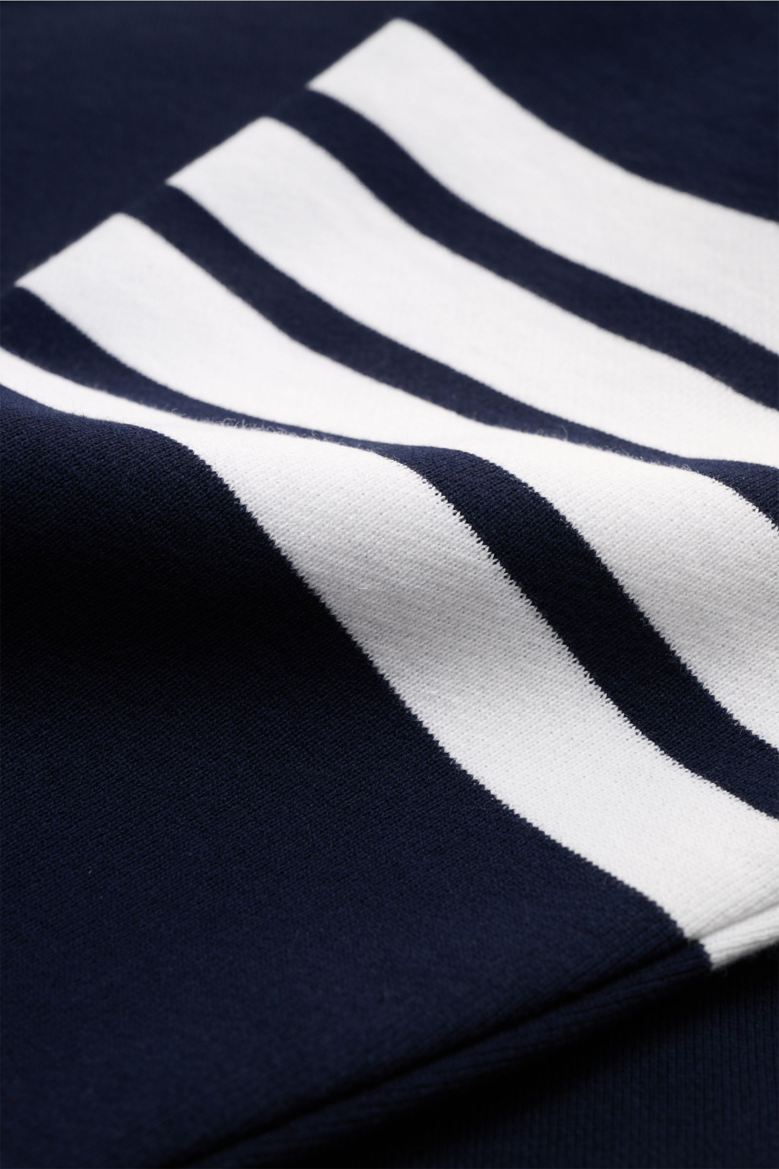Nahaufnahme des navyfarbenen Thom Browne Rundhals-Sweatshirts mit weißen Kontraststreifen am Ärmel, weiche Baumwolle, Grosgrain-Schlaufe rückseitig, aus schräger Perspektive.