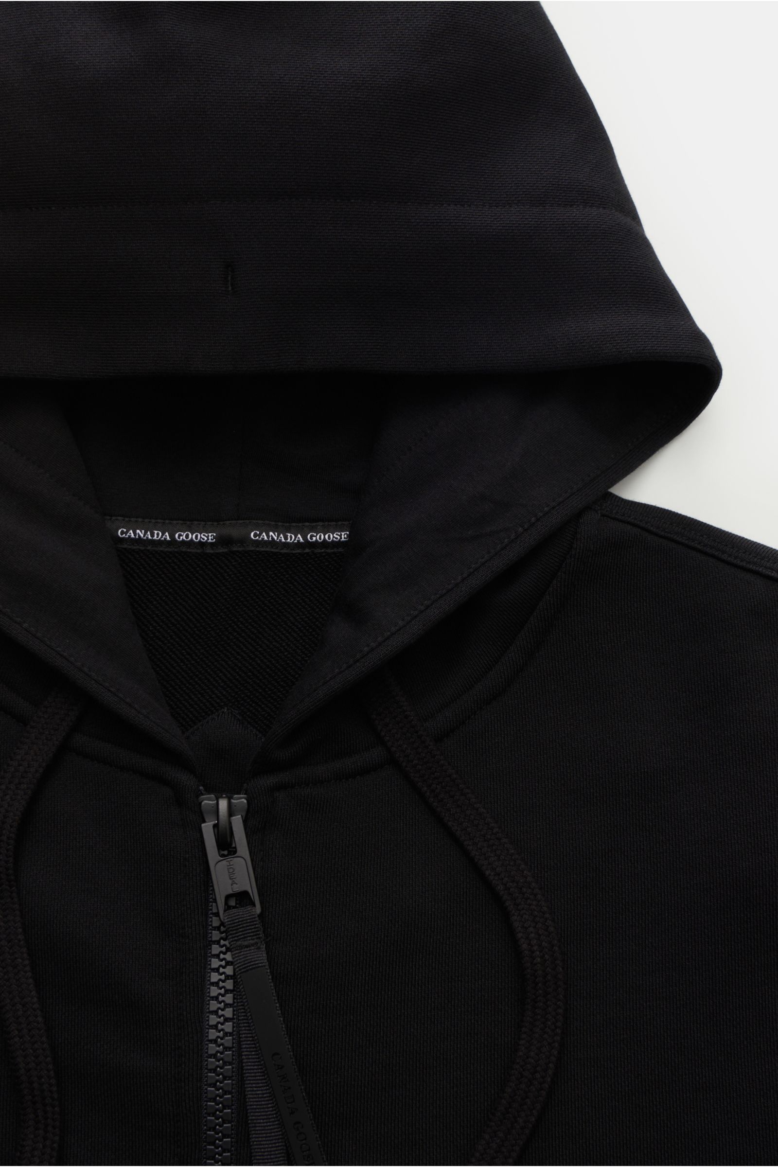 Close-up top-down view of the Canada Goose Sweatjacke 'Huron' schwarz black hoodie showing robust zipper, hood with drawstring, and CANADA GOOSE label. Sweat aus reiner Baumwolle, weicher Griff, Regular Fit, robuster Reißverschluss, Kapuze mit Tunnelzug, 