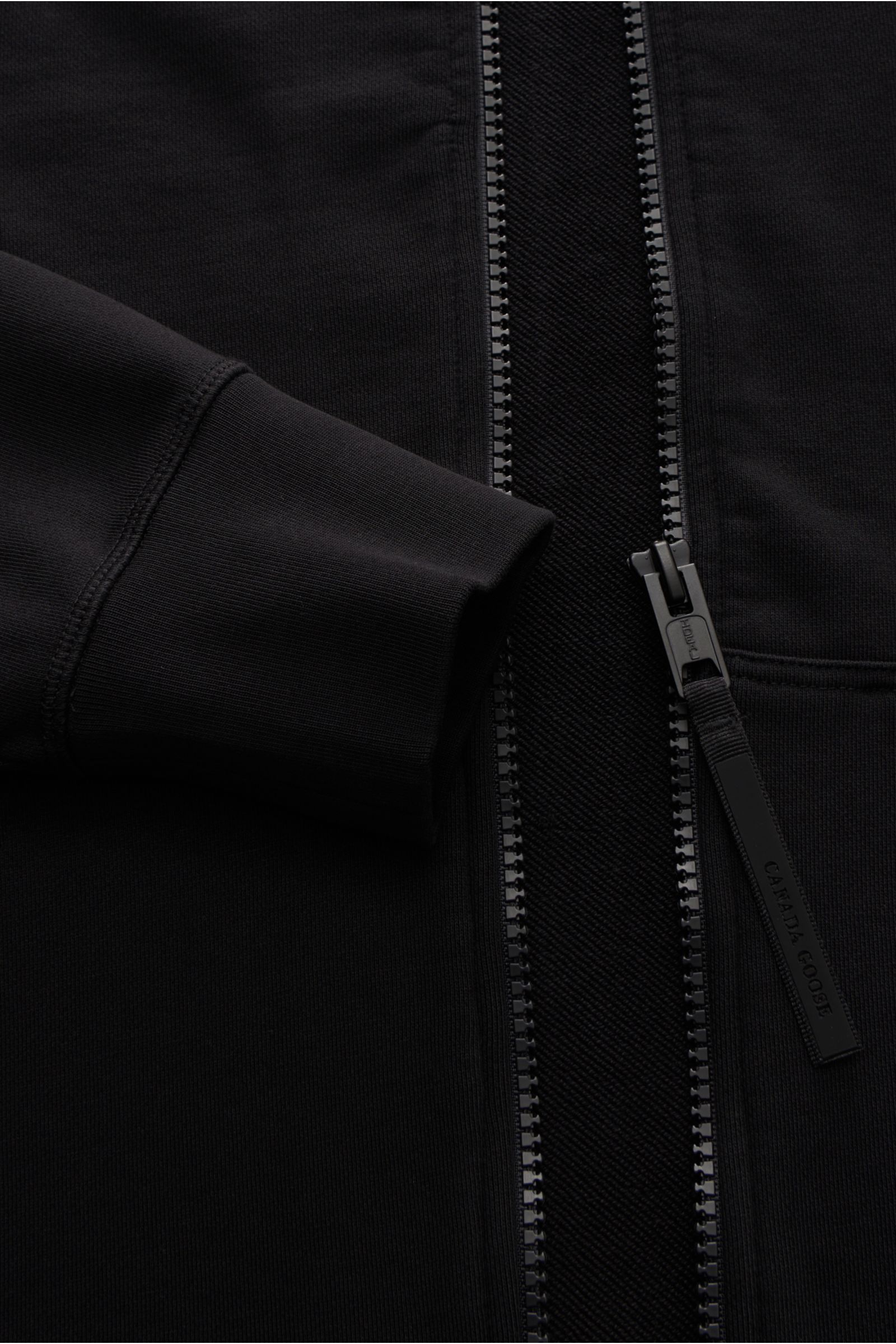 Close-up front view of the Canada Goose Sweatjacke 'Huron' schwarz, showing the robust zipper, ribbed cuff, and soft cotton fabric. Features: Sweat aus reiner Baumwolle, weicher Griff, Regular Fit, robuster Reißverschluss, Kapuze mit Tunnelzug, geteilte K