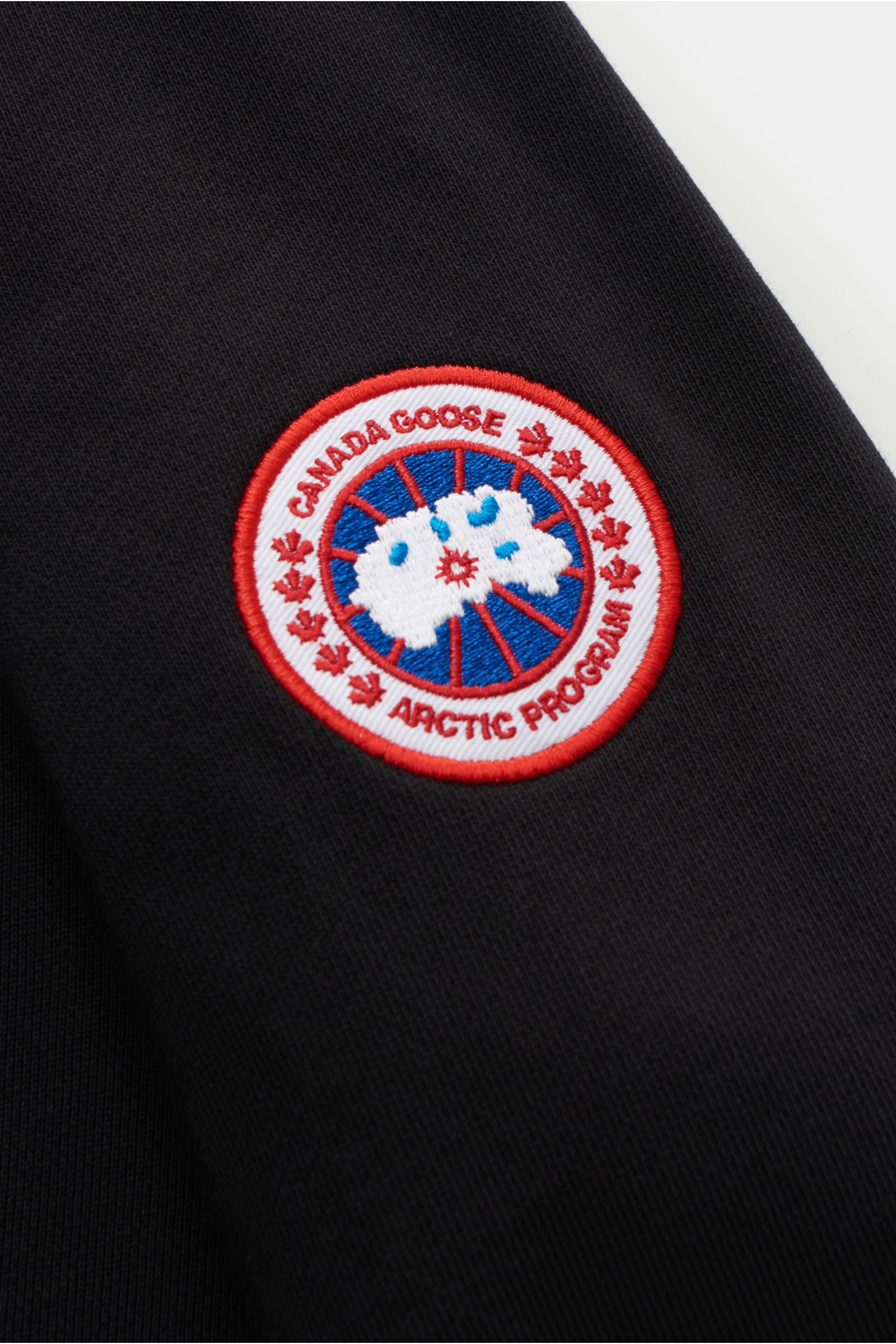 Close-up view of the CANADA GOOSE-Weblabel on the upper arm of the Canada Goose Sweatjacke 'Huron' schwarz, a sweat aus reiner Baumwolle with weicher Griff, Regular Fit, robuster Reißverschluss, Kapuze mit Tunnelzug, geteilte Kängurutasche, and komfortabl