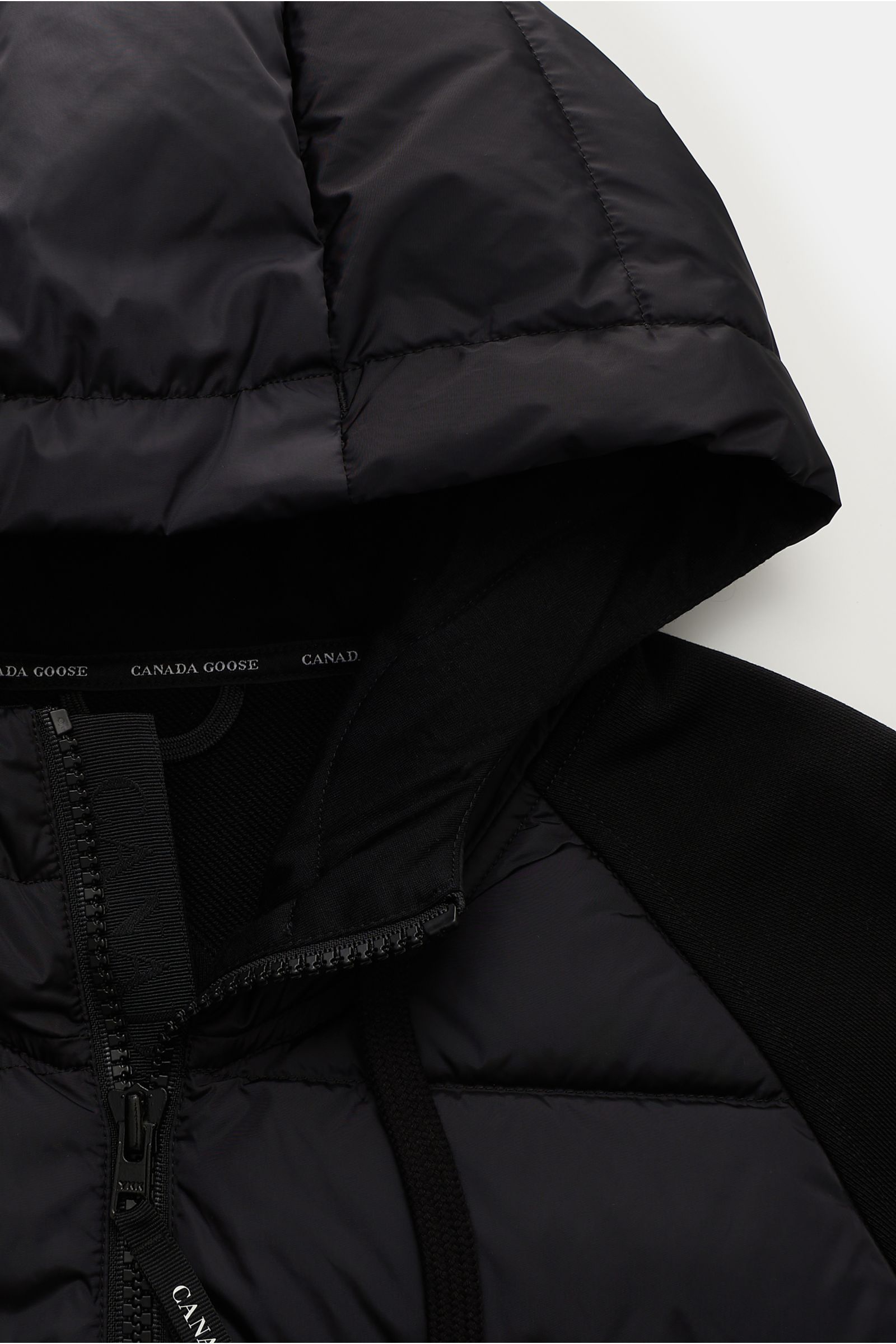 Close-up top-front view of the Canada Goose Sweatjacke 'HyBridge Huron' schwarz showing the black down-filled hood, cotton sweat fabric with soft texture, two-way zipper, and ribbed cuffs. CANADA GOOSE kombiniert bei der mittelschweren Hybrid-Kapuzenjacke
