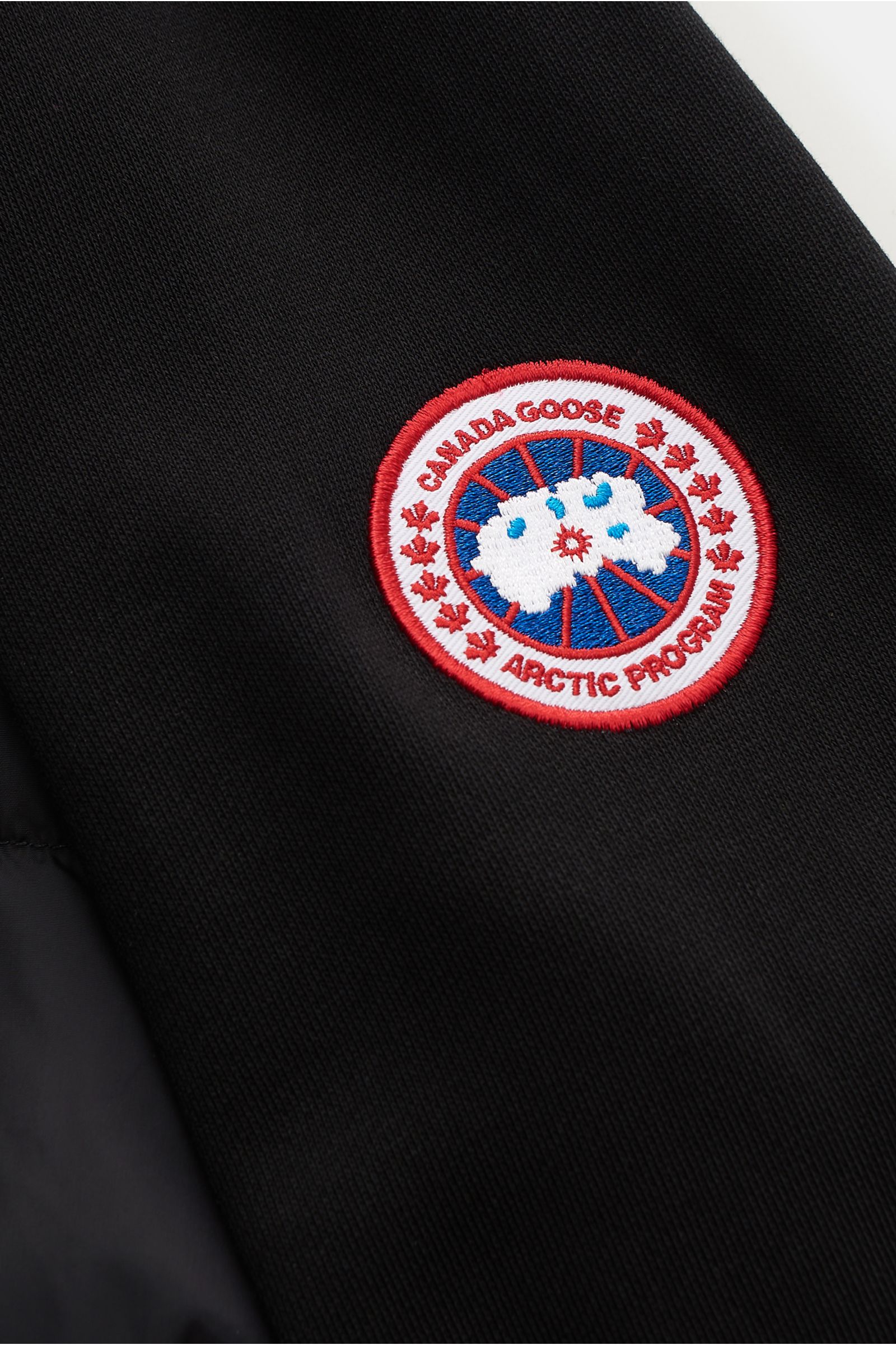 Close-up front-side view of the Canada Goose Sweatjacke 'HyBridge Huron' schwarz, showing the black soft cotton fabric and the embroidered Canada Goose Arctic Program patch on the upper arm. CANADA GOOSE kombiniert bei der mittelschweren Hybrid-Kapuzenjac