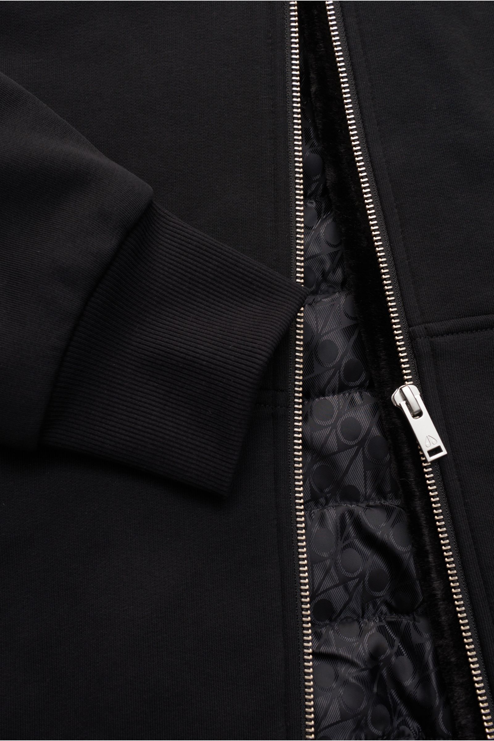 Close-up front view of the black Moose Knuckles Sweatjacke 'Classic Bunny' schwarz showing ribbed cuff, silver zipper, and patterned quilted lining.

Perfekt für kalte Tage: Dank der leichten Wattierung und einer Kapuze mit abnehmbarem Faux-Fur-Innenfu