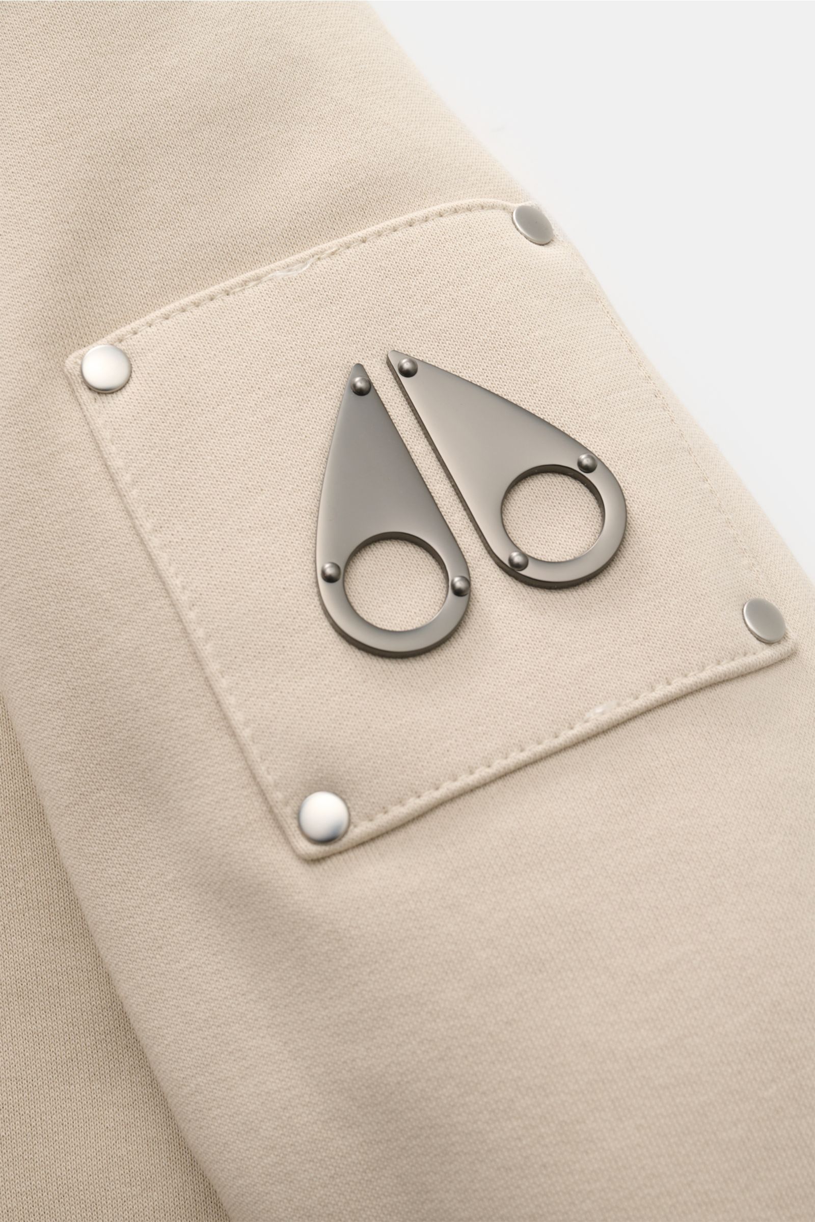 Close-up front view of the Moose Knuckles Sweatjacke 'Classic Bunny' creme showing the metal logo on a stitched fabric patch with rivets. Perfekt für kalte Tage: Dank der leichten Wattierung und einer Kapuze mit abnehmbarem Faux-Fur-Innenfutter sind Si