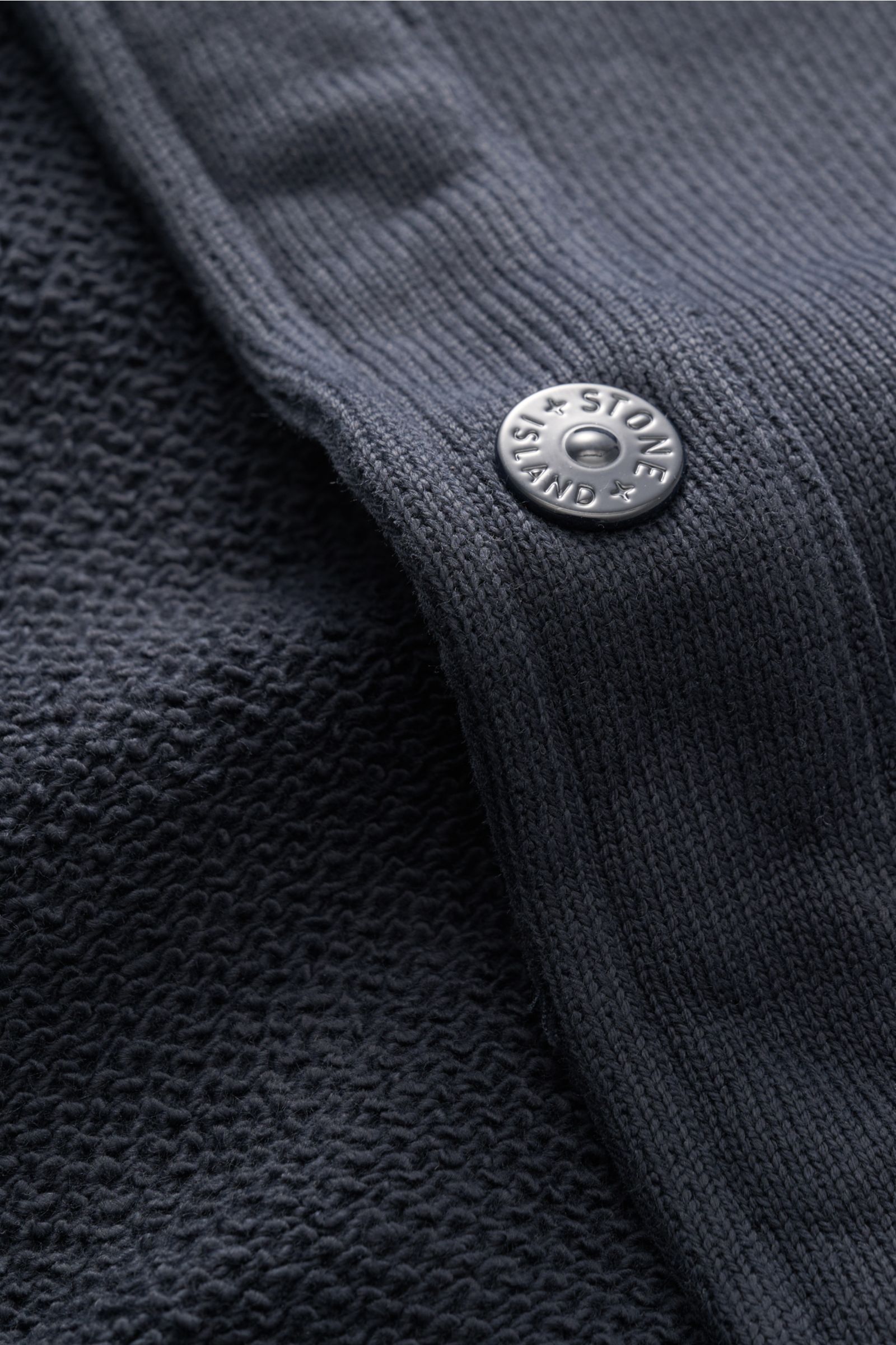 Nahaufnahme der Stone Island Sweatjacke 'Ghost' navy aus der 'Ghost' Capsule Collection, Fokus auf Knopf mit Logo und Struktur der reine Baumwolle, mittelfeines Strickbild, Regular Fit, Umlegekragen, aufgesetzte Brusttaschen mit Patte, Ärmelabschlüsse mit