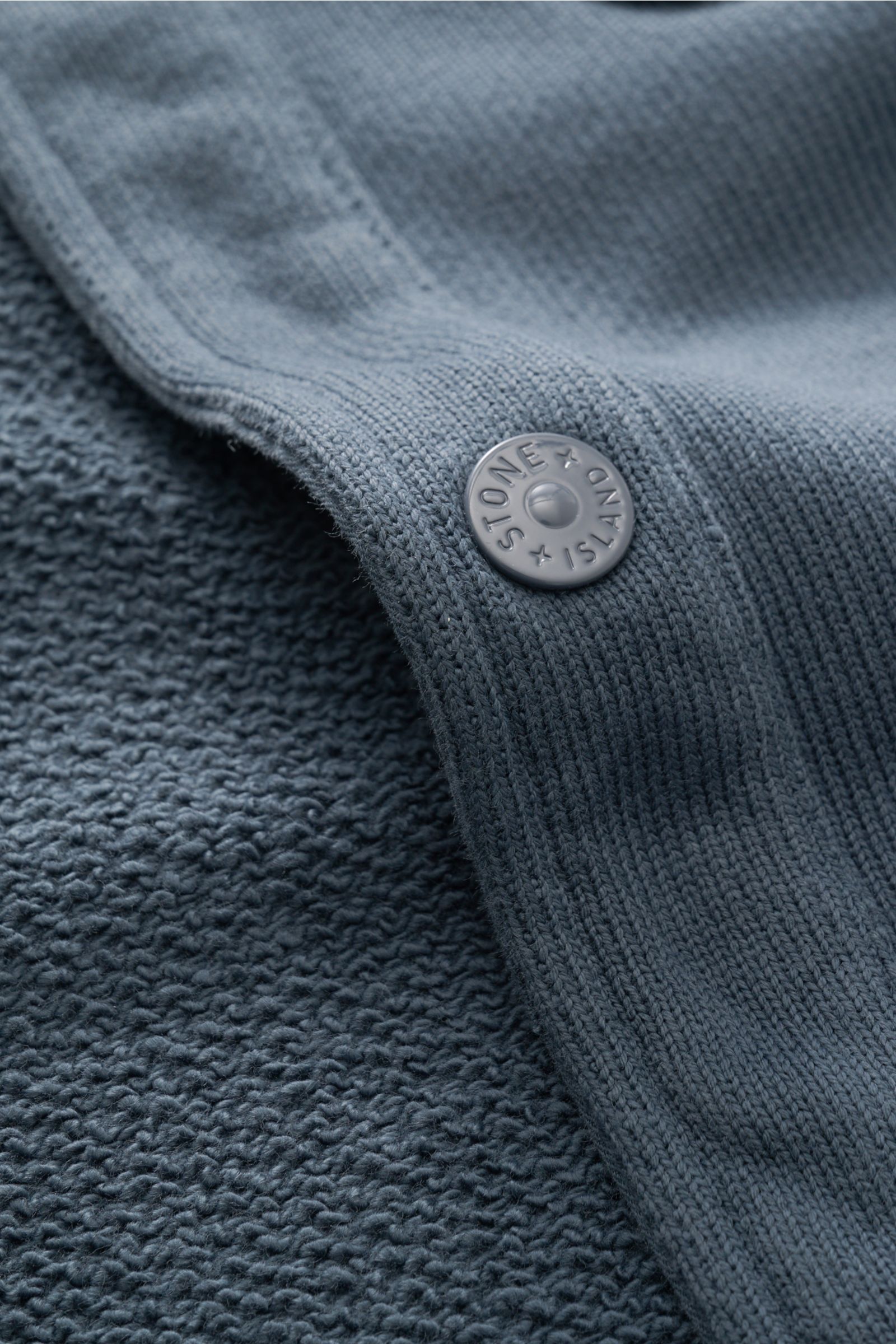 Nahaufnahme der Stone Island Sweatjacke 'Ghost' graublau aus der 'Ghost' Capsule Collection, Detail der reinen Baumwolle, mittelfeines Strickbild und Druckknopf mit Markenlogo, Perspektive seitlich oben.
