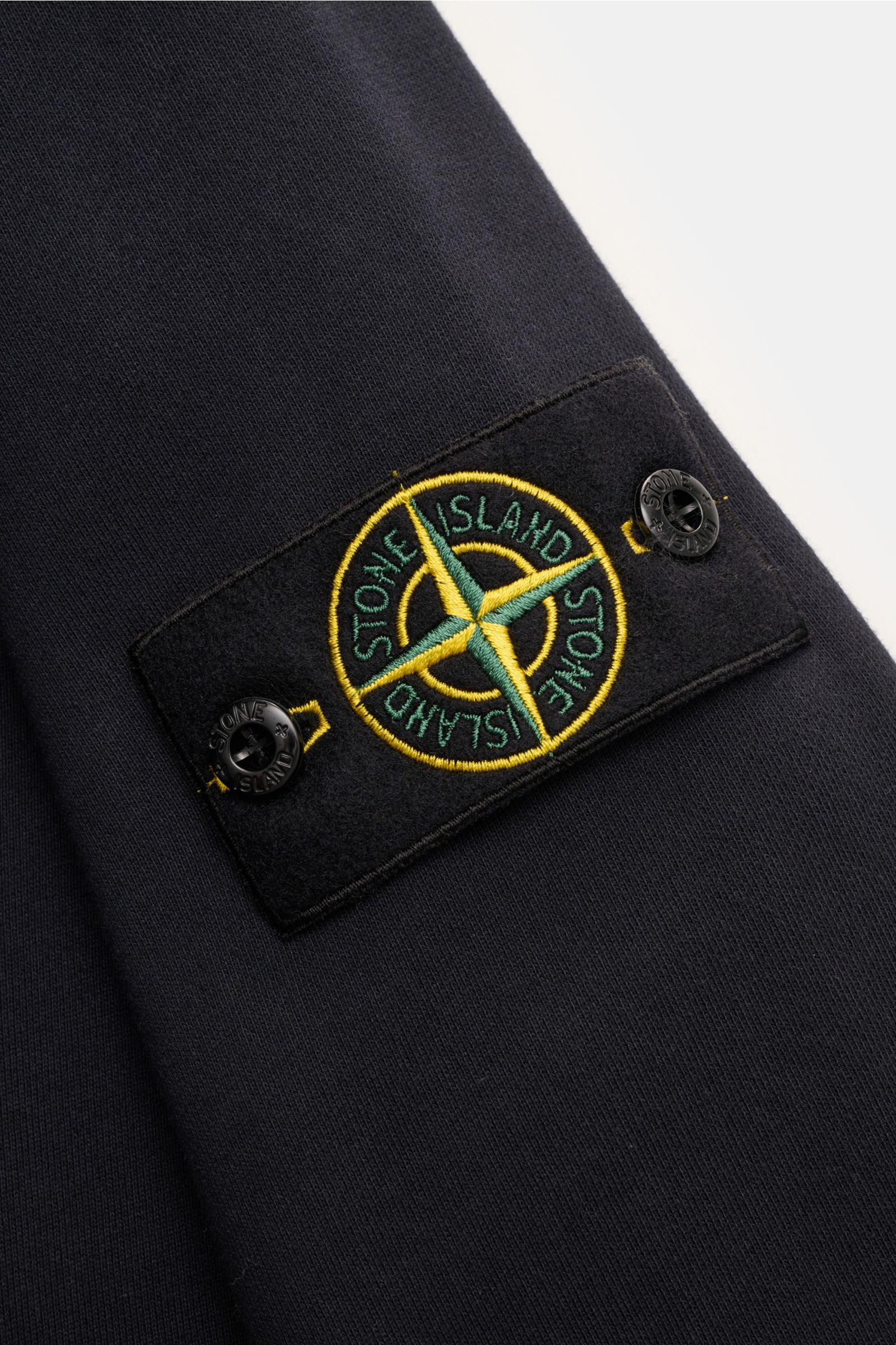 Nahaufnahme der Stone Island Sweatjacke dark navy, Fokus auf das Kompass-Badge an der Ärmelaußenseite, Baumwolle, Rippbündchen.