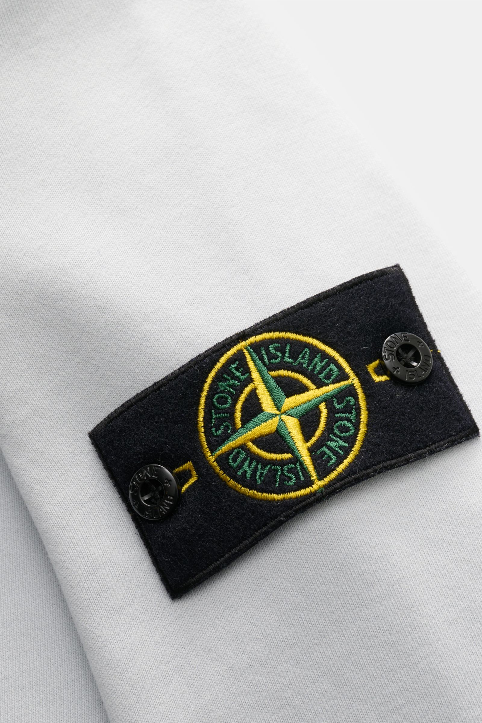 Stone Island Sweatjacke hellgrau, Nahaufnahme des typischen Kompass-Badges auf weichem Baumwollstoff aus schräger Perspektive.