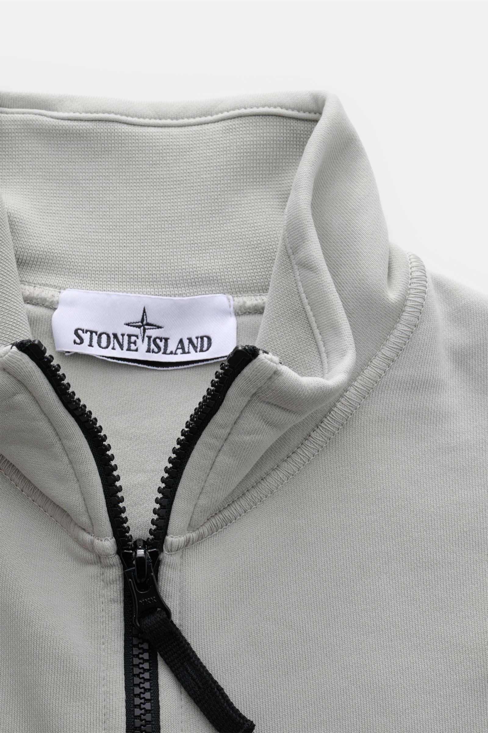 Close-up front view of Stone Island Sweatjacke hellgrau showing soft cotton fabric, robust black zipper, and high stand-up collar with Stone Island label.

Description: Entspanntes Basic von STONE ISLAND: Mit sportiver Lässigkeit begleitet uns die stüc