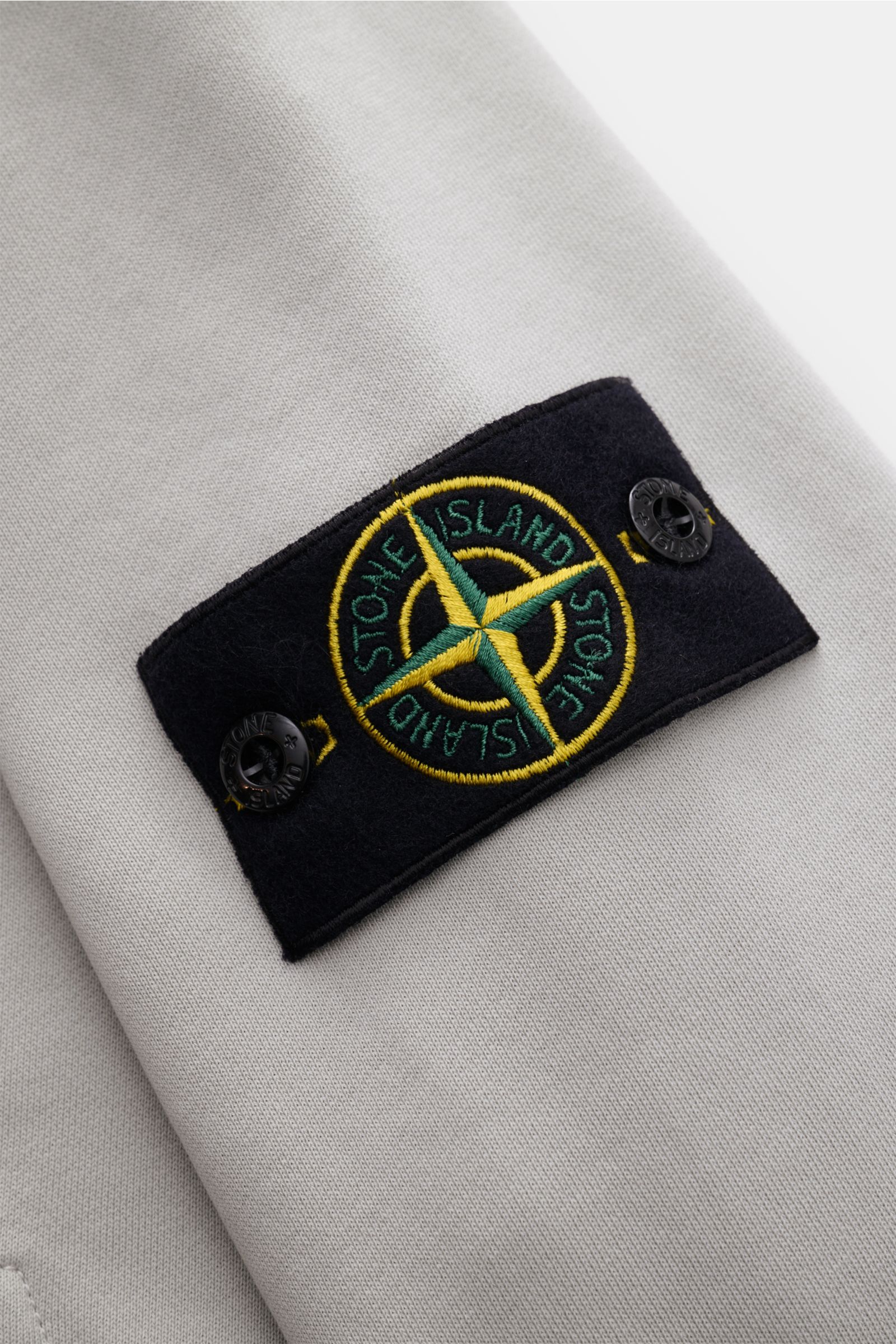 Close-up view of the Stone Island Sweatjacke hellgrau showing the garment dyed soft cotton fabric and the typical compass badge on the sleeve, captured from a top angle. Entspanntes Basic von STONE ISLAND: Mit sportiver Lässigkeit begleitet uns die stü