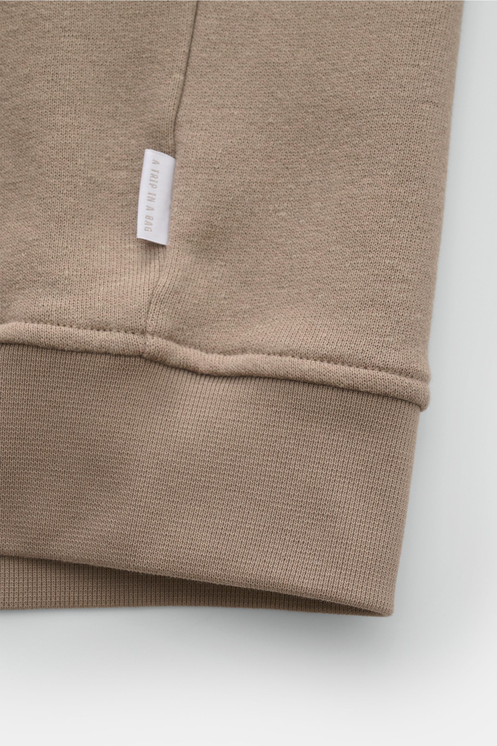 Detailaufnahme der 04651/ A trip in a bag Sweatjacke 'CL Bomber' taupe, Perspektive auf das flexible, gerippte Bündchen und Weblabel, aus Bio-Baumwolle und Leinen, garment-dyed mit weichem Griff.