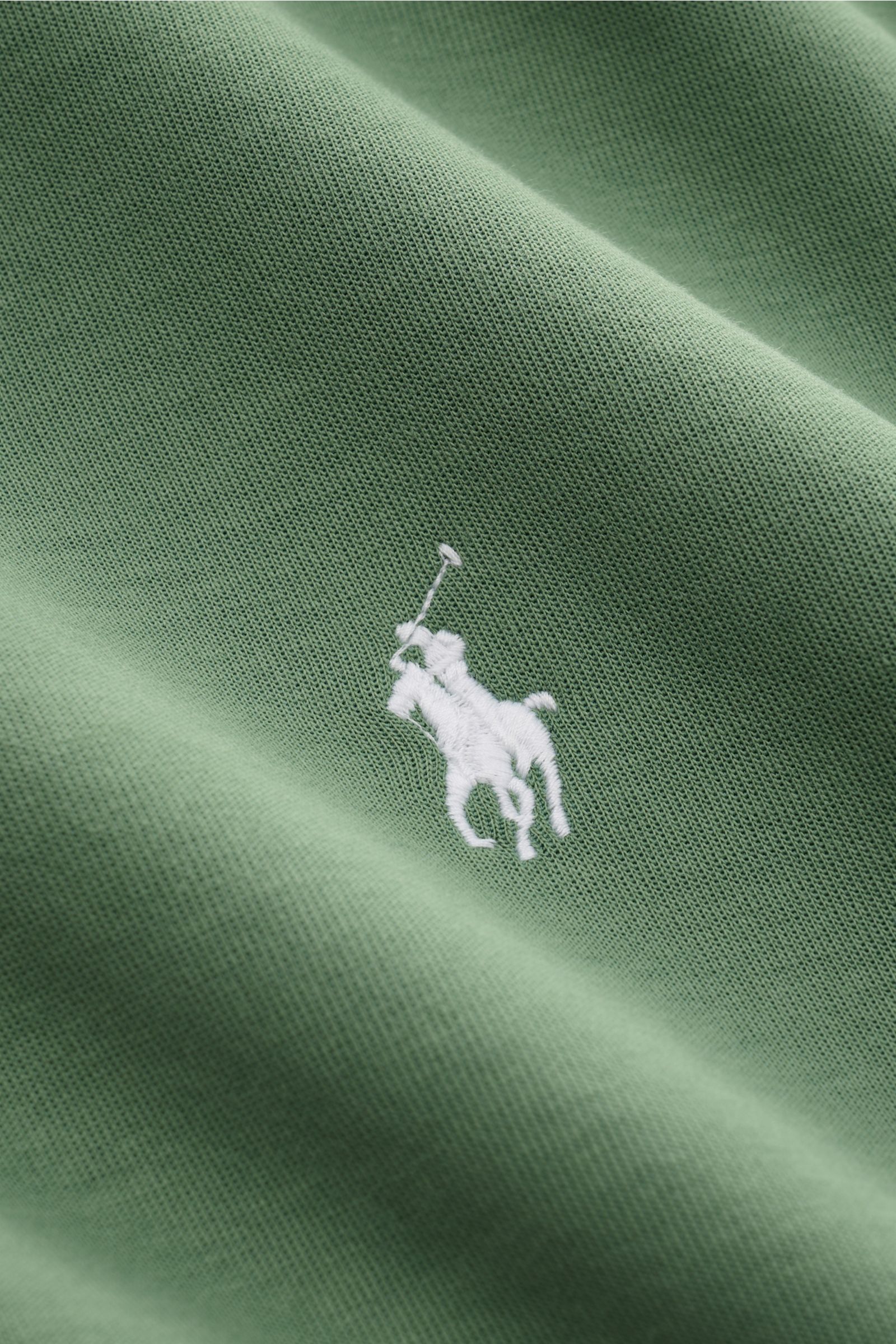 Green fabric close-up showing the white signature logo stitching of the Polo Ralph Lauren Hooded jumper green, photographed from an angled top view. Jersey made of cotton and microfibre, soft feel, regular fit, drawstring hood, kangaroo pocket, signature 