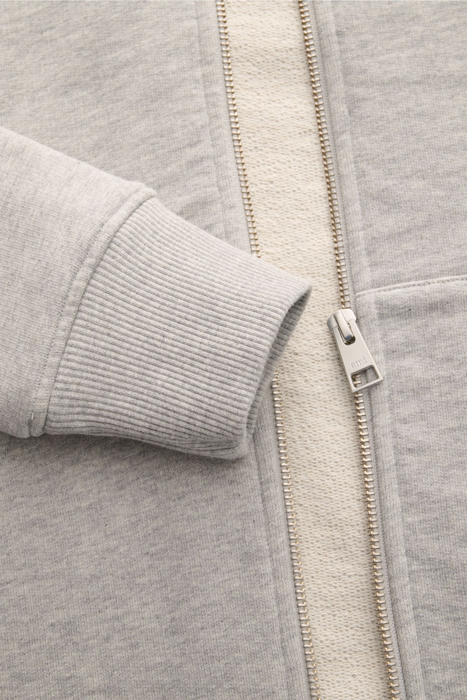 Nahaufnahme der Ami Paris Sweatjacke hellgrau meliert von schräg oben, Details wie Rippbündchen, Metallreißverschluss und Herz-Stitching sichtbar.