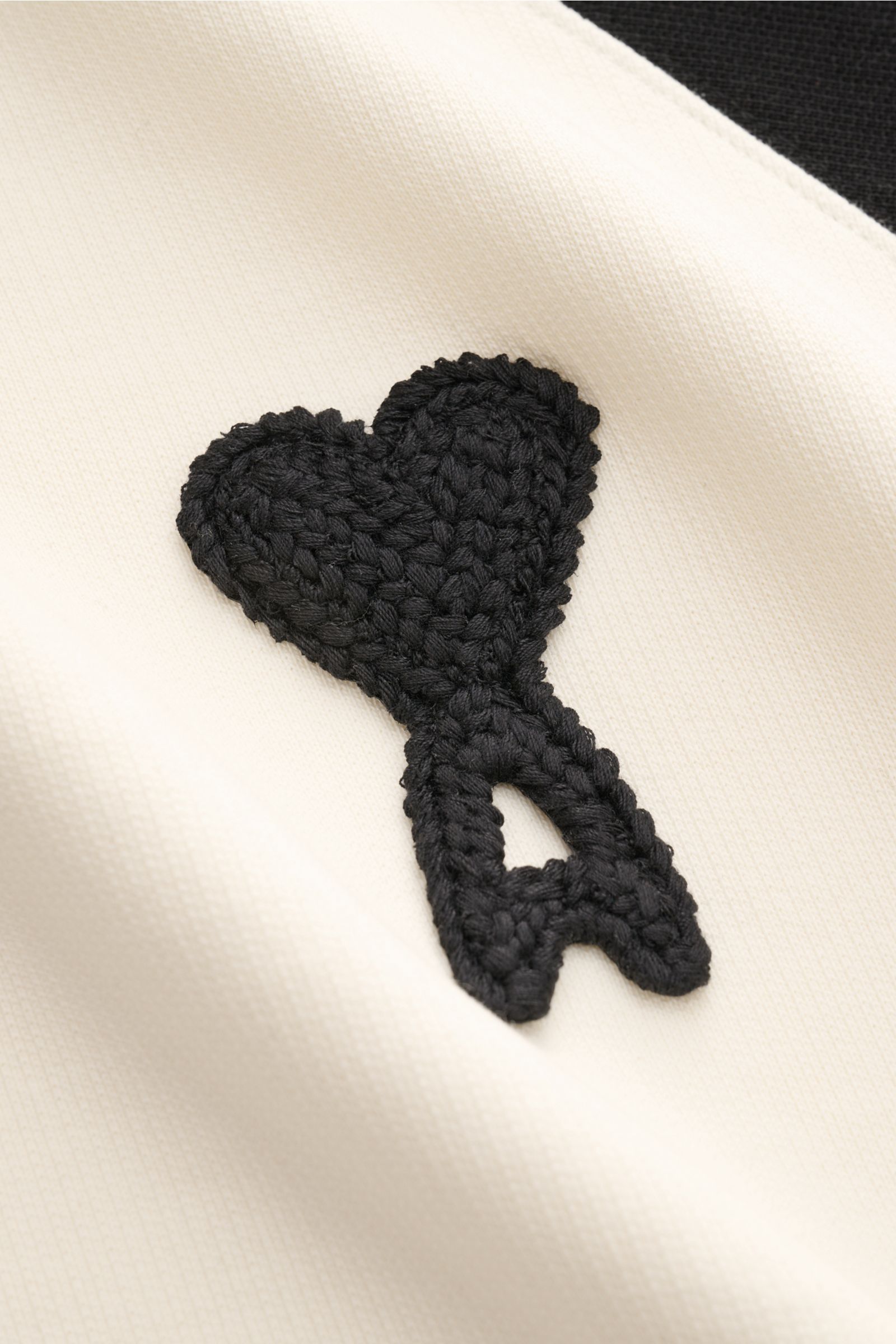 Nahaufnahme des Ami Paris Kapuzenpullovers 'Boxy Fit' creme/schwarz mit schwarzem Herz-Logo-Stitching, reine Baumwolle, Raglanärmel.