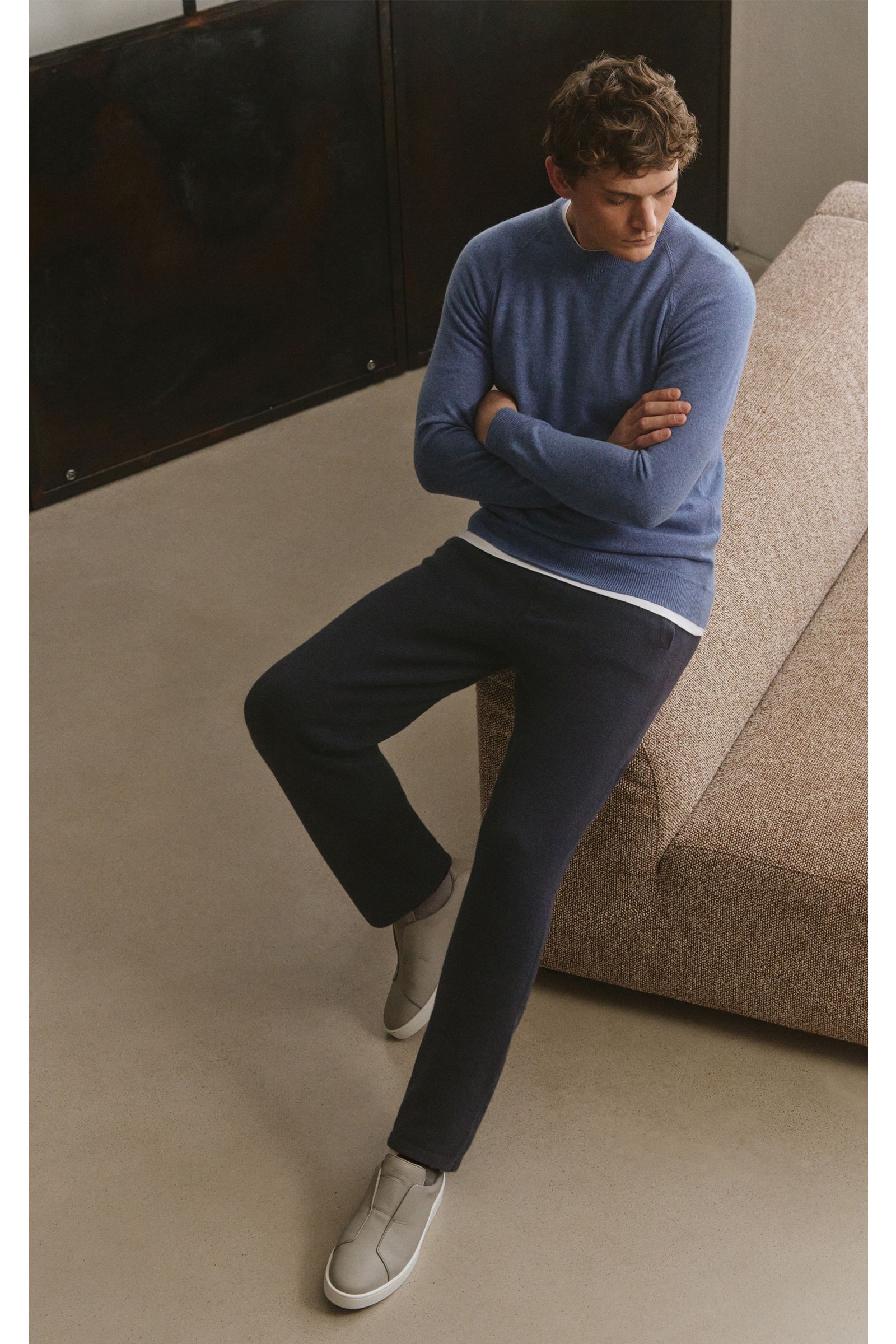 Mann sitzt seitlich auf Sofa, trägt von Braun Cashmere Jogpants navy mit geradem Schnitt, elastischem Bund, Hüft- und Leistentaschen, fotografiert von oben.