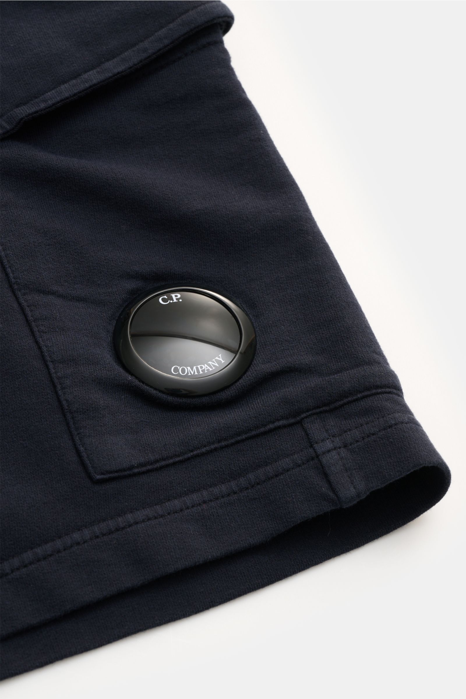 C.P. Company Cargo-Sweatshorts dark navy, Nahaufnahme der Cargo-Tasche mit Linsen-Detail, weicher Baumwollsweat, elastischer Bund, Slim Fit, leichte sommerliche Qualität, Perspektive von oben.