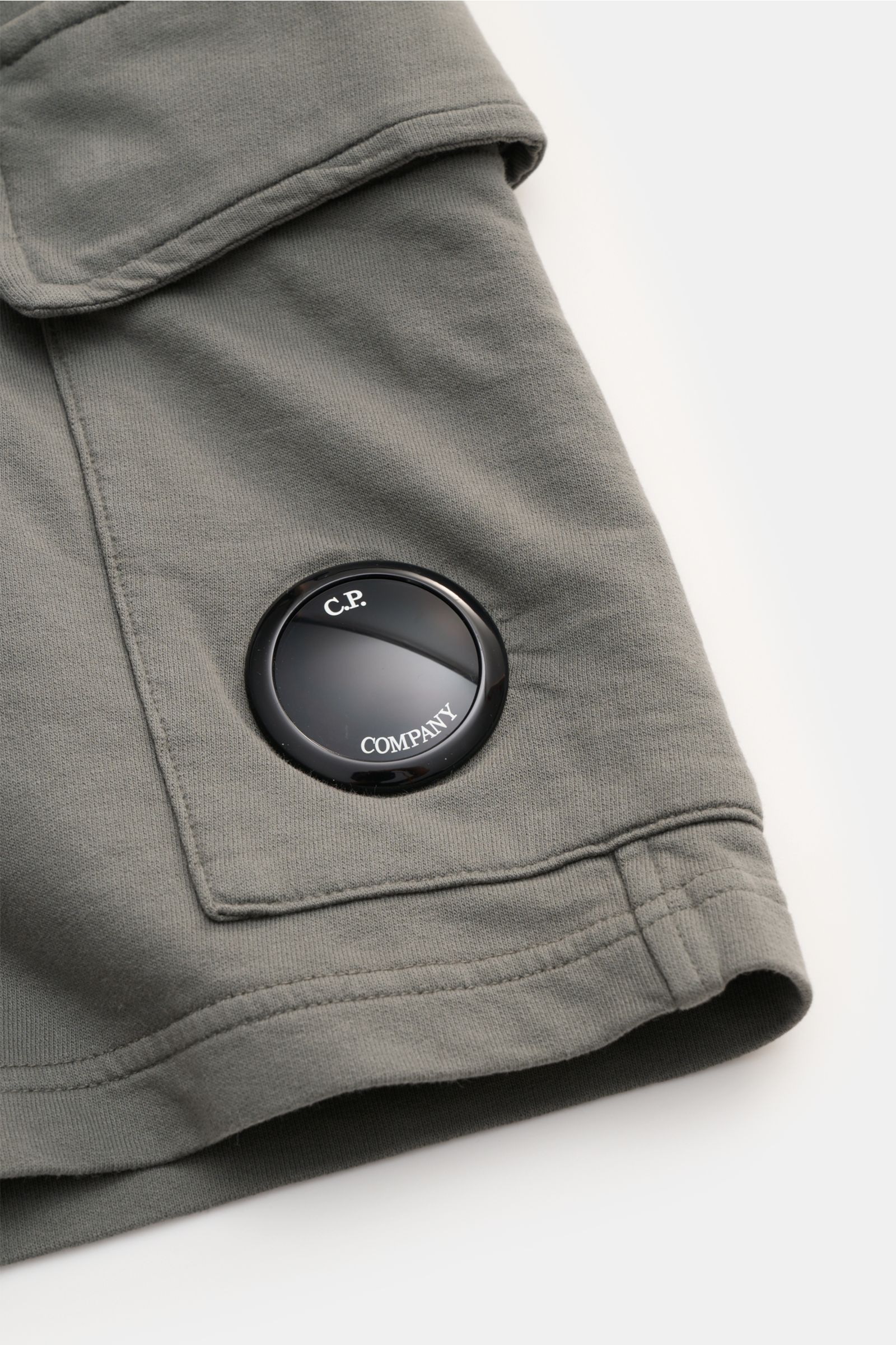 Nahaufnahme der C.P. Company Cargo-Sweatshorts graugrün von schräg oben, zeigt weiches Baumwoll-Sweatmaterial, Cargo-Tasche mit Linsen-Detail, elastischen Bund, Slim Fit, leichte Qualität, garment-dyed, angedeuteter Eingriff und Taschen in der Seitennaht.