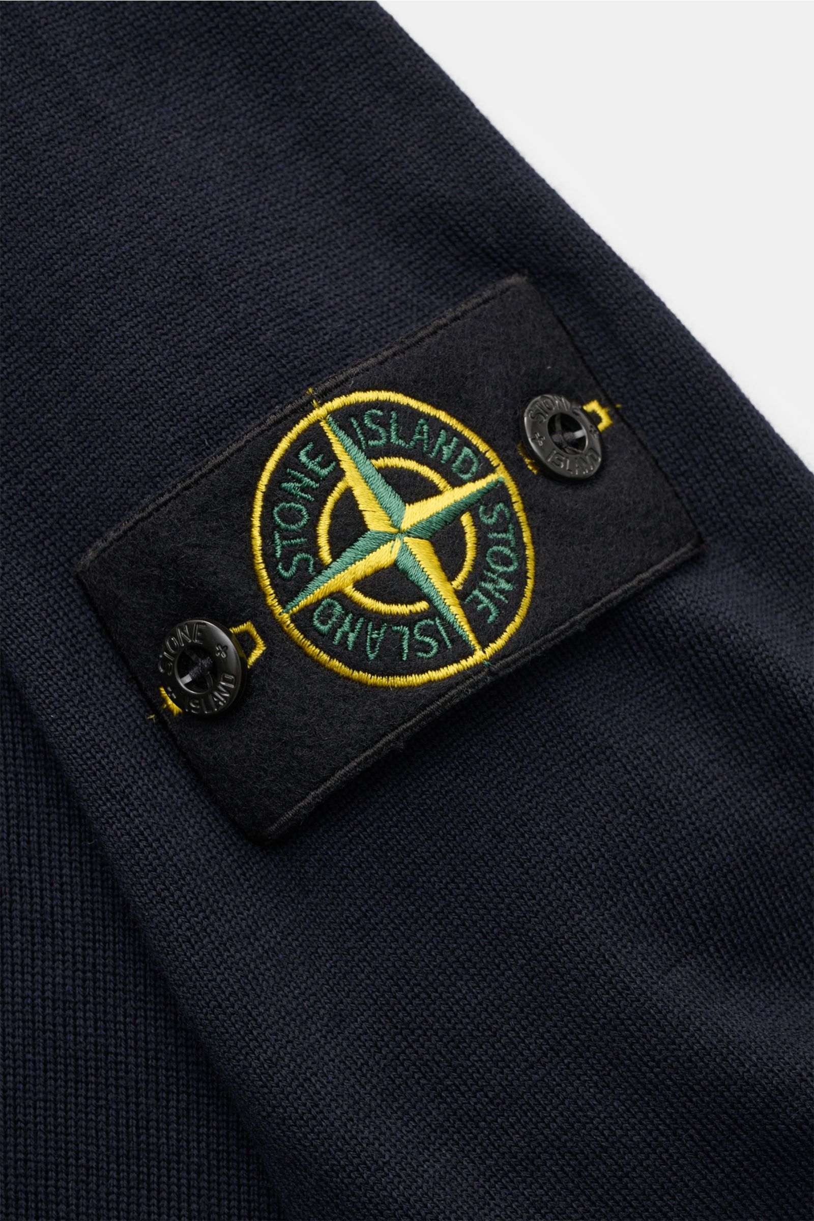 Close-up of the sleeve of Stone Island Troyer 'Light Pure Wool' navy, showing the brand’s compass badge in yellow and green on black felt, photographed from a slightly angled top view. A knit classic with timeless appeal: This Troyer from STONE ISLAND fea