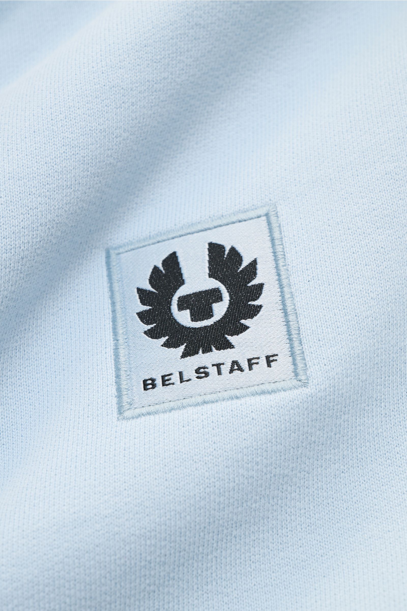 Nahaufnahme des hellblauen Belstaff Sweat-Troyer 'Quarter Zip' mit weichem Griff und sichtbarem BELSTAFF-Weblabel auf der Brust.