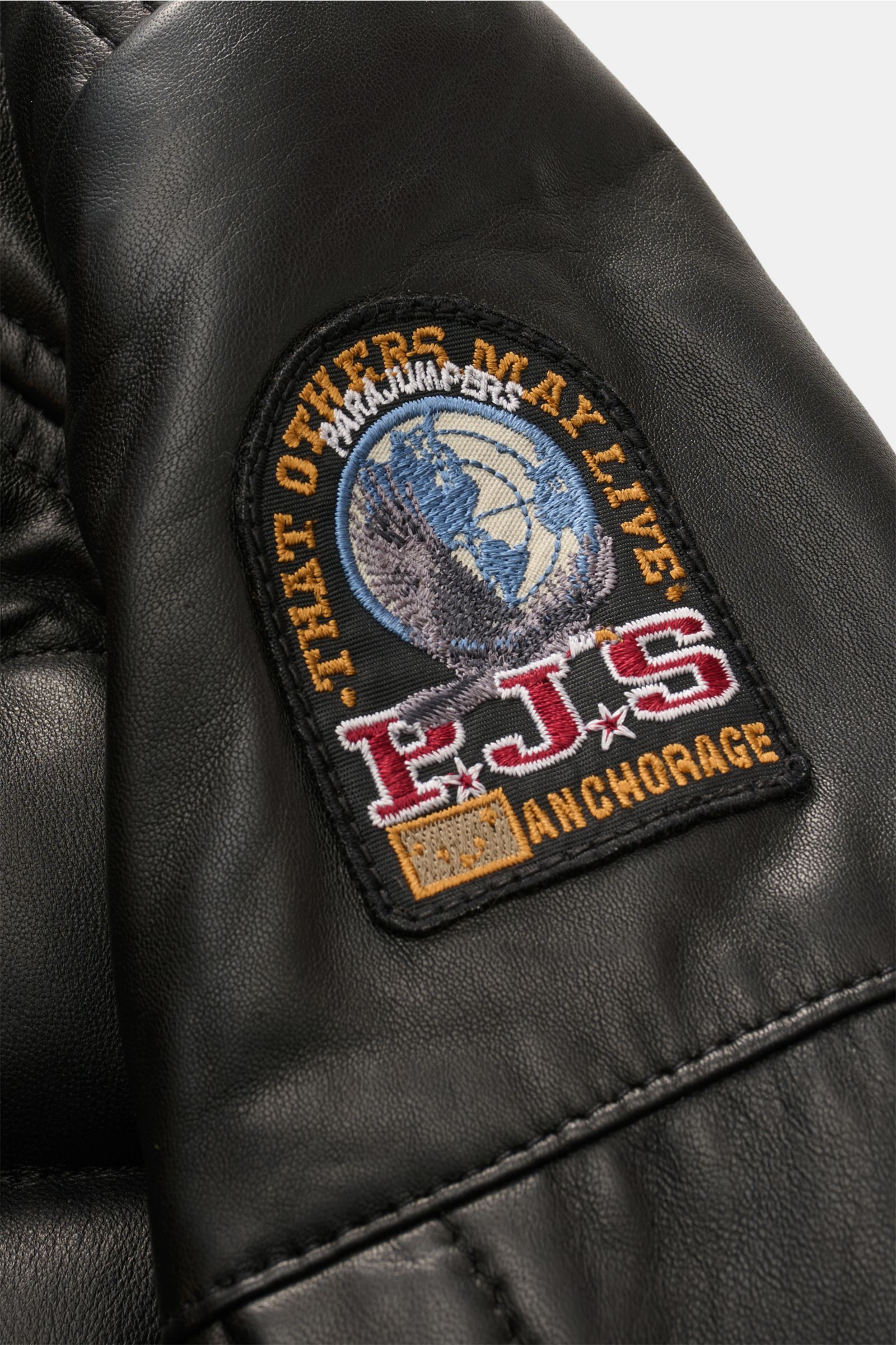 Close-up side view of the Parajumpers Lederjacke 'Ernie' schwarz showing the detailed embroidered PJS patch on smooth black lamb leather with stitched seams, highlighting its butter-soft material and craftsmanship. Ein Hauch Abenteuer, verpackt in pure