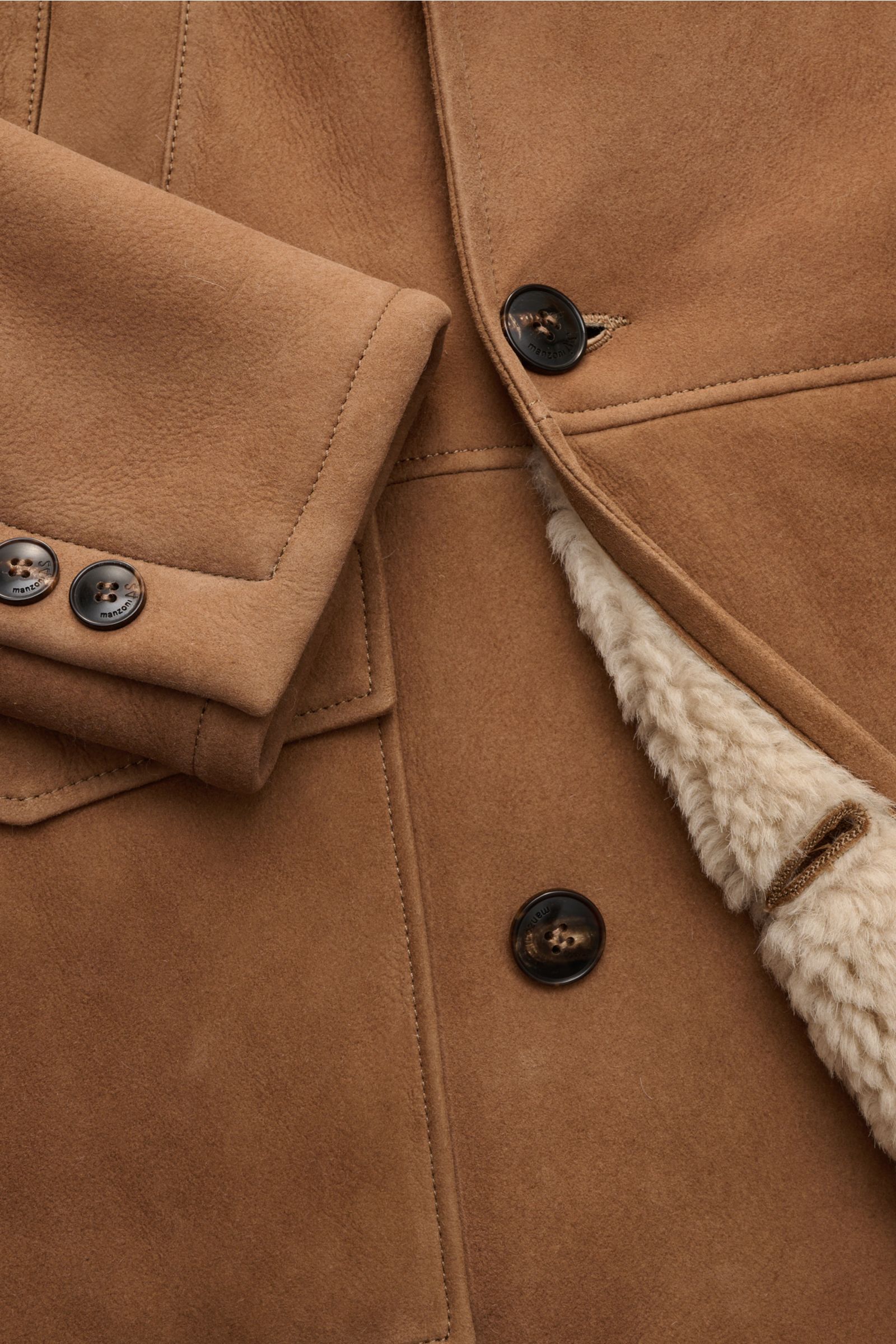Nahaufnahme des Manzoni 24 Lammfellmantels braun aus reinem Lammveloursleder, zeigt zweireihige Drei-Knopf-Leiste, Spitzrevers, weiches Fellfutter, Handwärmetaschen, Hüfttaschen mit Patte und Ärmelabschlüsse mit Knöpfen, fotografiert von oben.