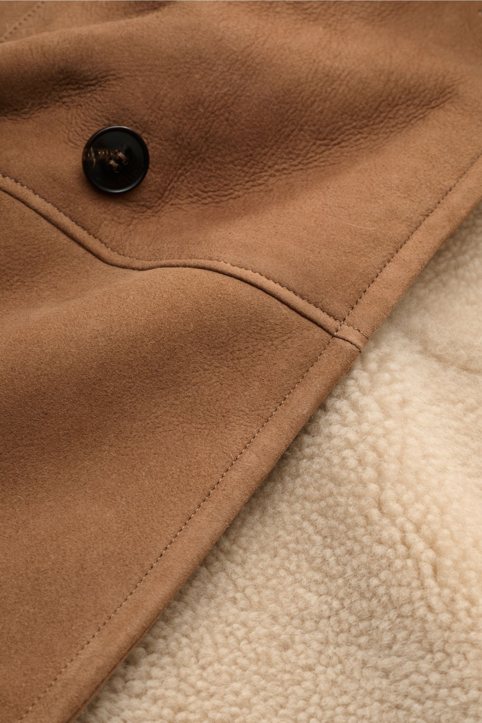 Nahaufnahme des Manzoni 24 Lammfellmantels braun, Detailansicht der weichen Lammveloursleder-Außenseite mit Knopf und cremefarbener Lammfell-Innenseite.