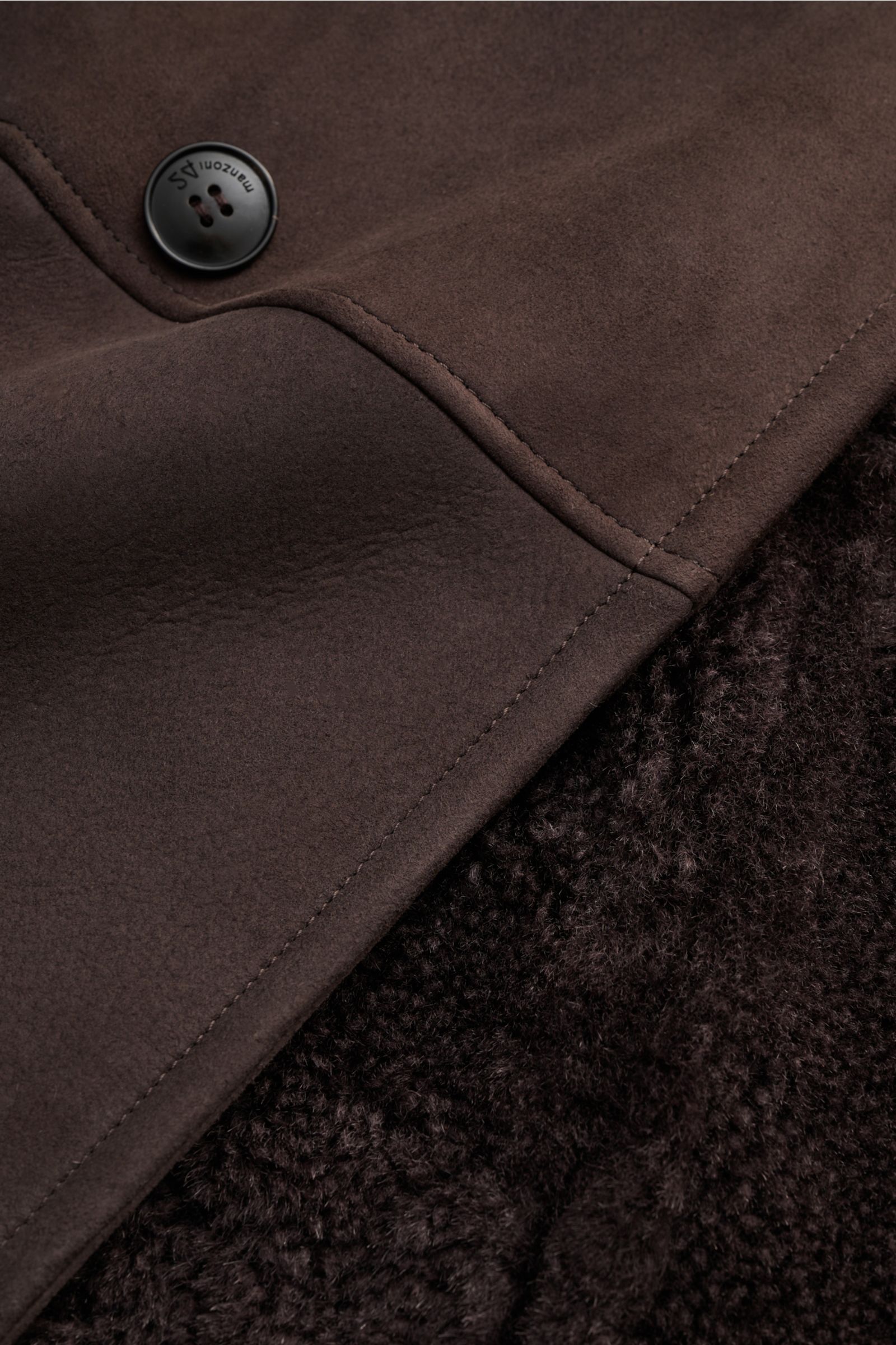 Nahaufnahme des Manzoni 24 Lammfellmantels dunkelbraun von schräg oben, zeigt feines Lammveloursleder außen, weiche Innenseite aus Lammfell, Knopf mit Logo, Reverskragen, Slim Fit, Taschen und Details wie Ärmelknöpfe und Rückenmittelschlitz.