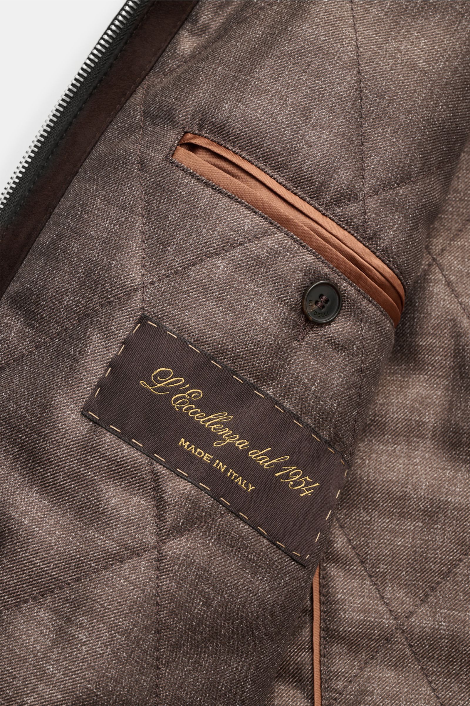 Close-up interior view of the Mandelli Veloursleder-Blouson dunkelbraun showing quilted lining, brown piping, buttoned inner pocket, and a label with gold text 'L’Eccellenza dal 1954 Made in Italy.' Luxuriöser Komfort für die Übergangszeit: Der Veloursled