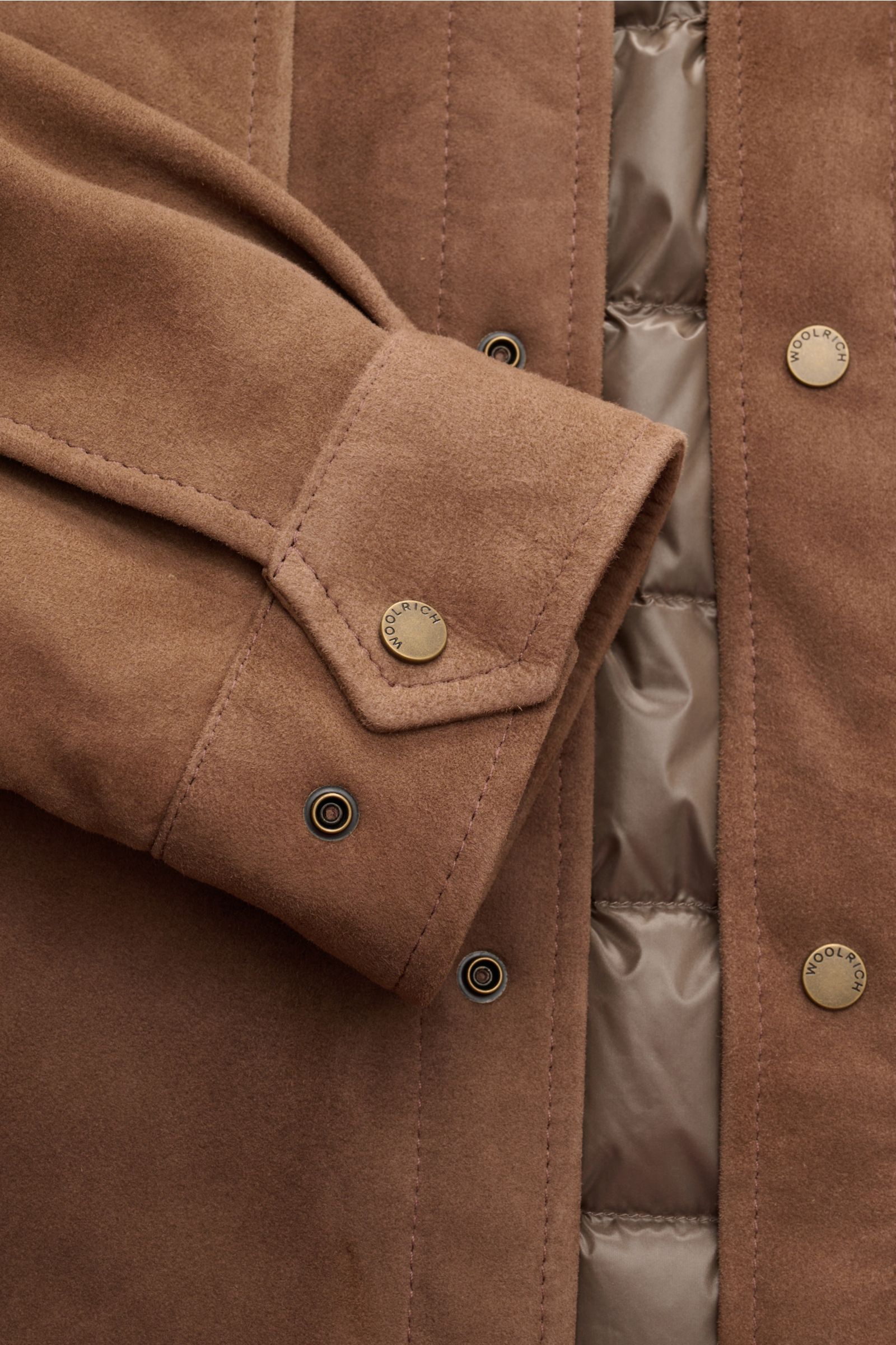 Nahaufnahme der Woolrich Veloursleder-Daunenjacke braun mit softem Ziegenvelours, leichter Daunenfüllung, Druckknopfleiste und gesteppter Innenseite.