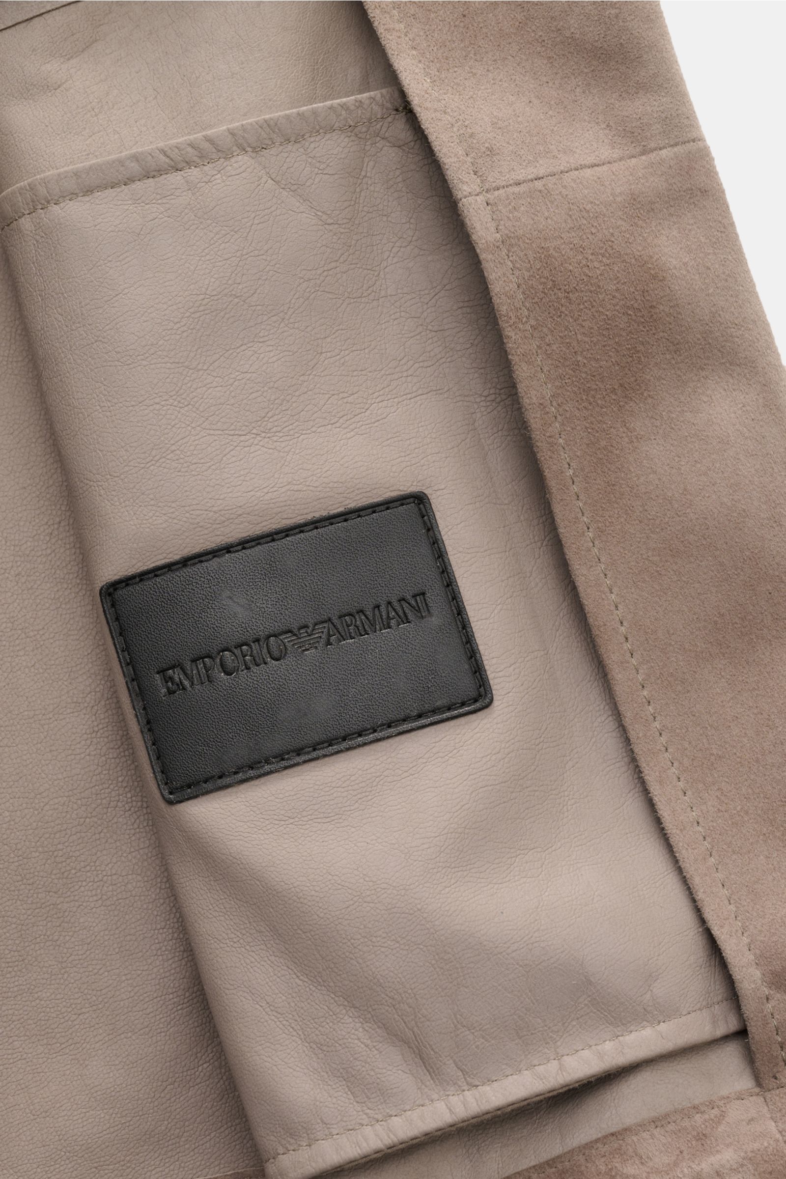 Close-up interior view of the Emporio Armani Velourslederjacke taupe showing soft goat leather, suede texture, and black embossed logo patch on lining. Urbaner Allrounder: Die Velourslederjacke von EMPORIO ARMANI kombiniert legeren Stil mit praktischer Ka