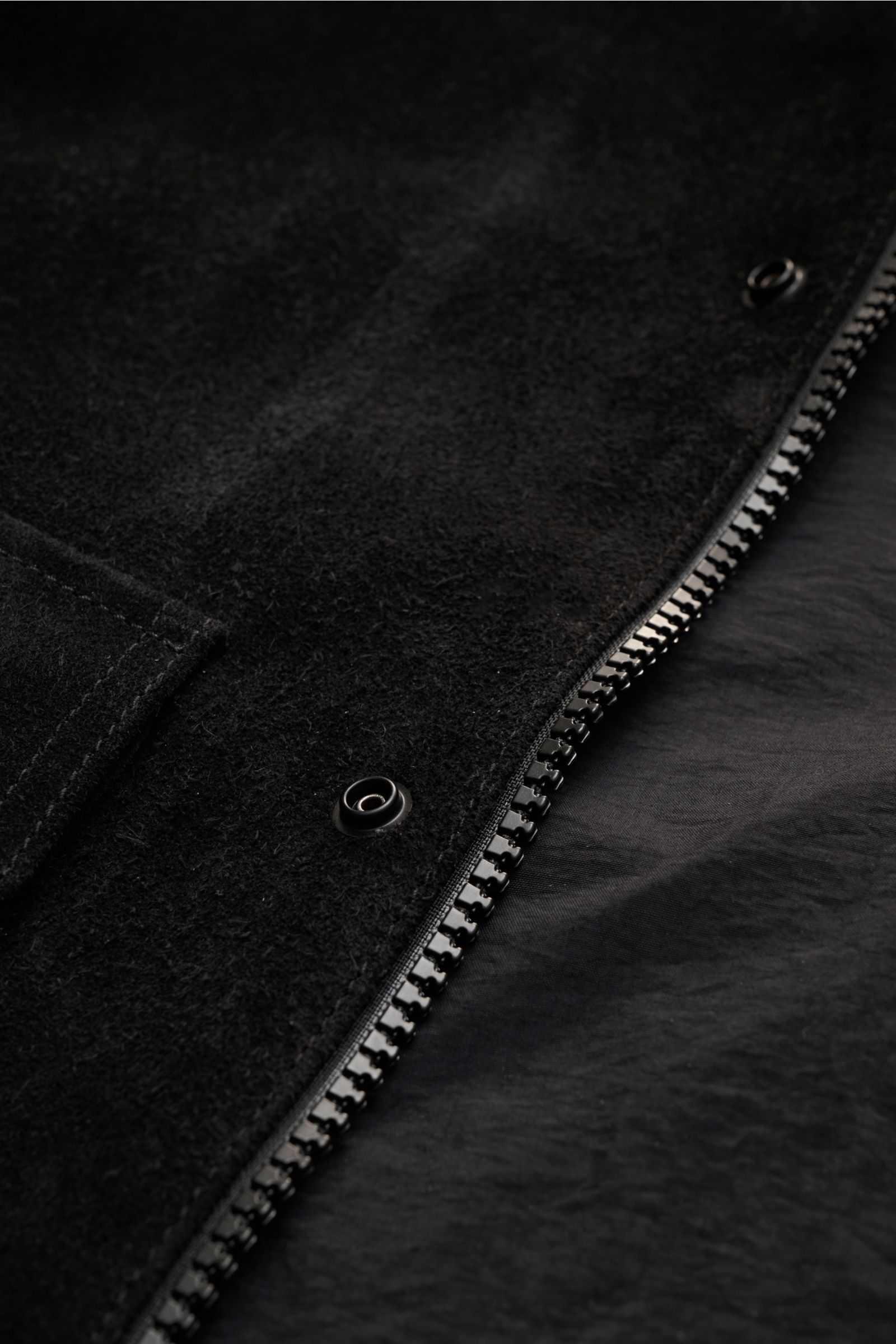 Nahaufnahme der Stone Island Velourslederjacke 'Ghost Piece' schwarz, Detailansicht des Zwei-Wege-Reißverschlusses, geschmeidiges Rindsleder mit weichem Griff und Windblende mit Druckknöpfen.