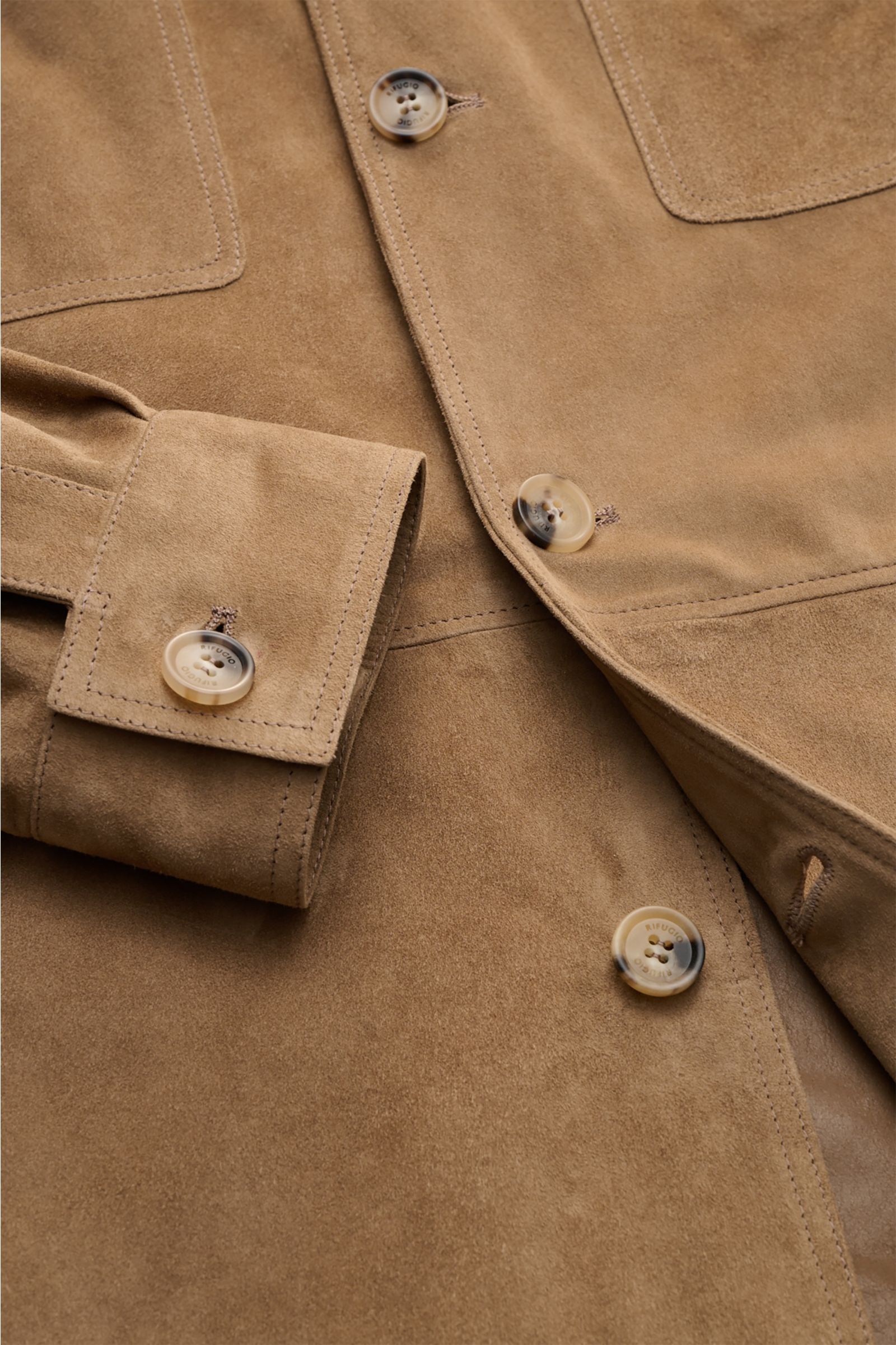 Nahaufnahme aus schräger Perspektive der Rifugio Velourslederjacke hellbraun, zeigt samtiges Veloursleder, Slim Fit, Knopfleiste, Umlegekragen, aufgesetzte Brusttaschen, Seitennahttaschen und Manschettenärmel im unconstructed Design.