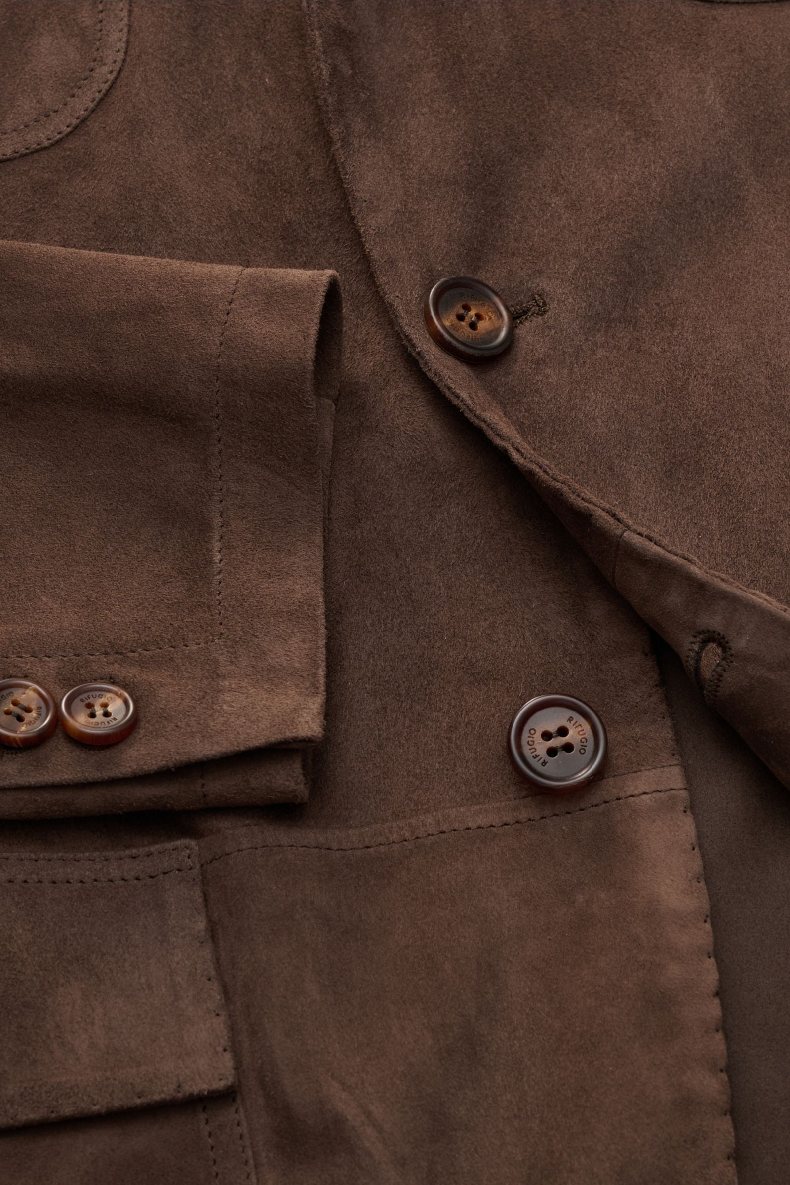 Close-up front view of Rifugio Suede jacket dark brown, showcasing supple suede, soft feel, slim fit, step lapel, fitted three-button style, four front pockets, buttoned cuffs, unconstructed design, and detailed stitching. The luxurious reinterpretation o