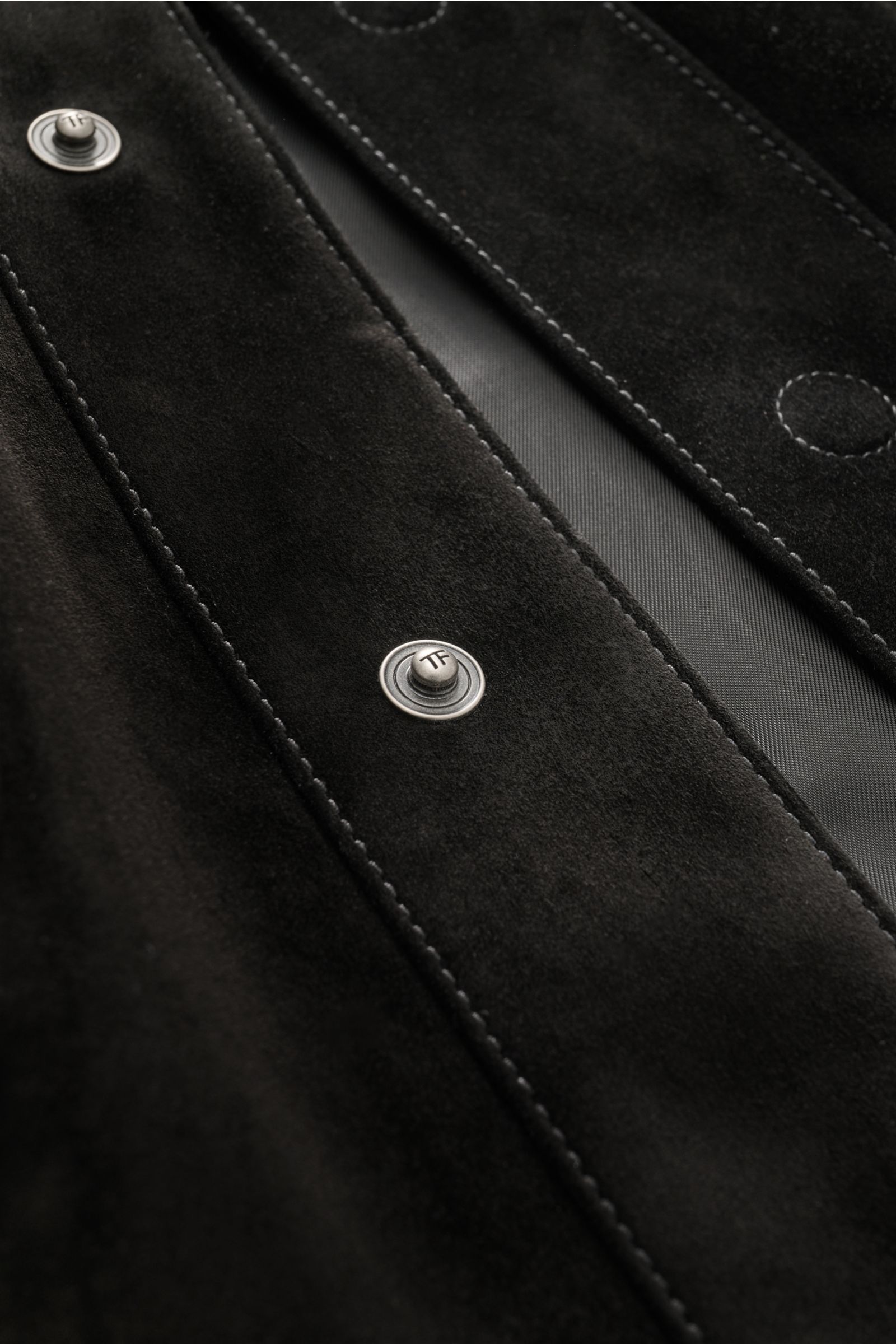 Close-up view of the Tom Ford Velourslederjacke schwarz showing fine black suede leather, detailed stitching, and branded metal snap buttons, photographed from a top angle. 
