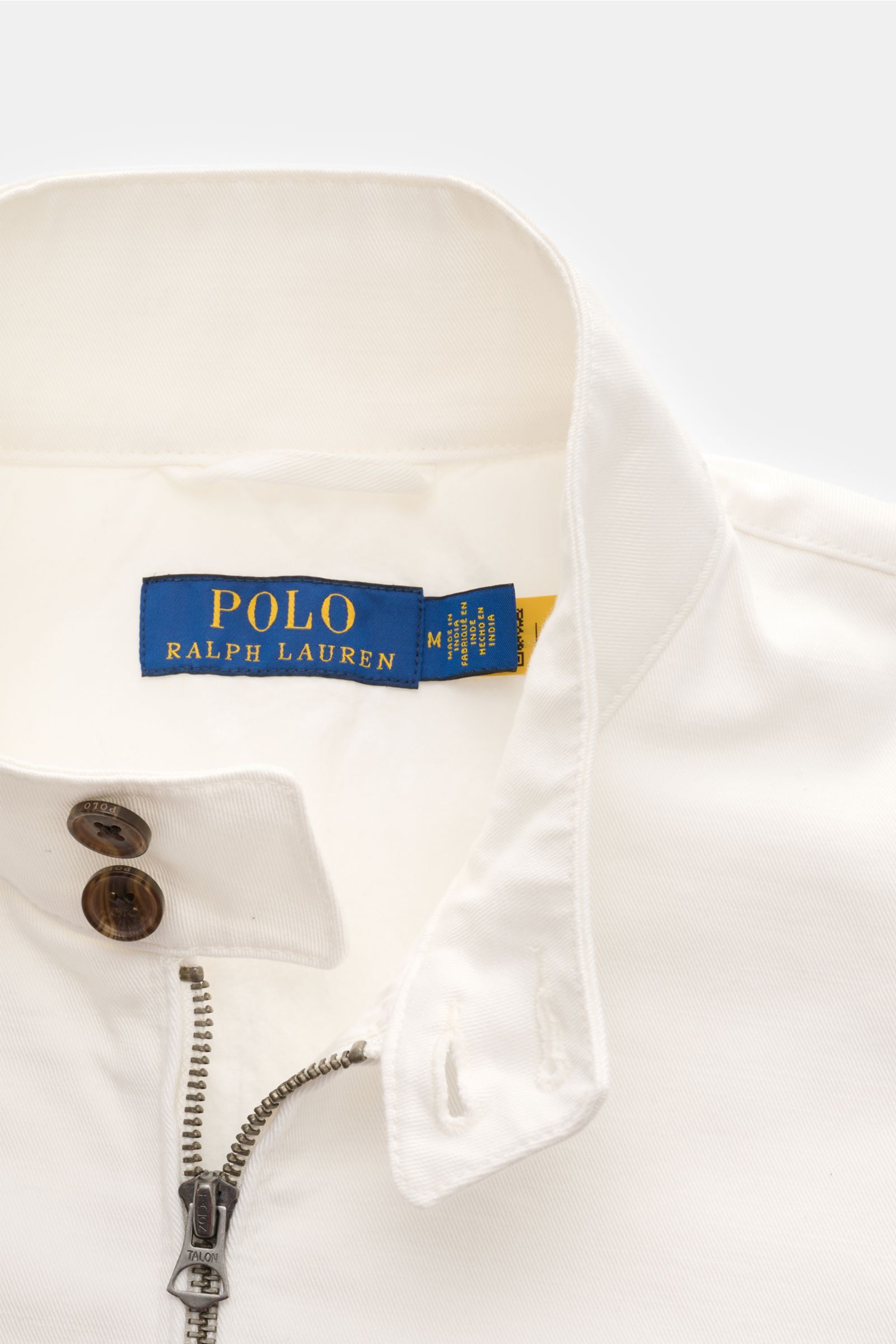 Nahaufnahme des weißen Polo Ralph Lauren Blousons aus Baumwolltwill mit Stehkragen, robustem Reißverschluss und Knöpfen, von oben fotografiert.