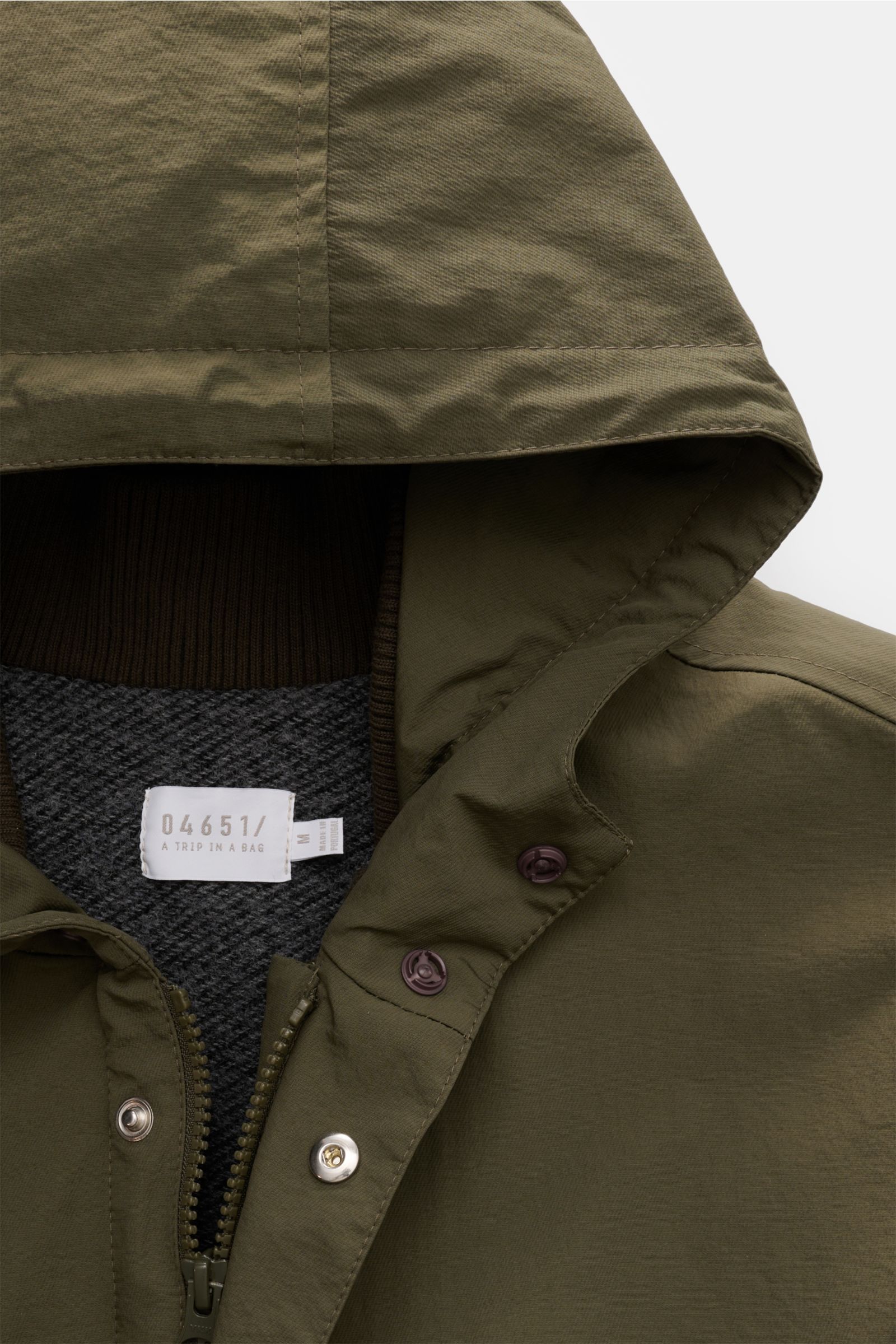 Close-up front view of the olive hood and upper chest area of the 04651/ A trip in a bag Parka 'The Triple' oliv, showing the high-quality microfiber fabric with a lightly textured feel, zipper, snap buttons on the wind flap, and the label inside the coll