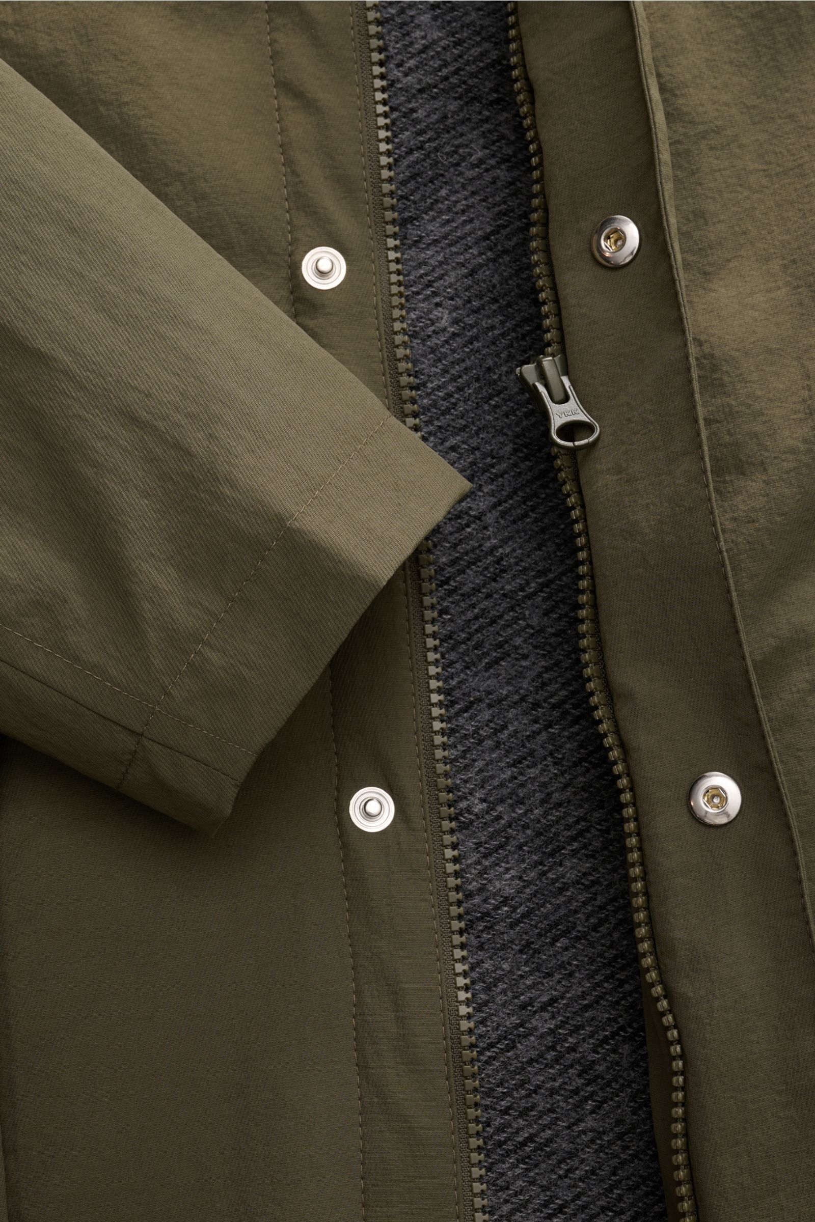 Close-up front view of the 04651/ A trip in a bag Parka 'The Triple' oliv showing olive microfiber fabric, zipper, snap buttons, and removable gray textured vest lining. Sportlich-leger durch die Übergangszeit: Der vielseitige Parka mit herausnehmbarer We