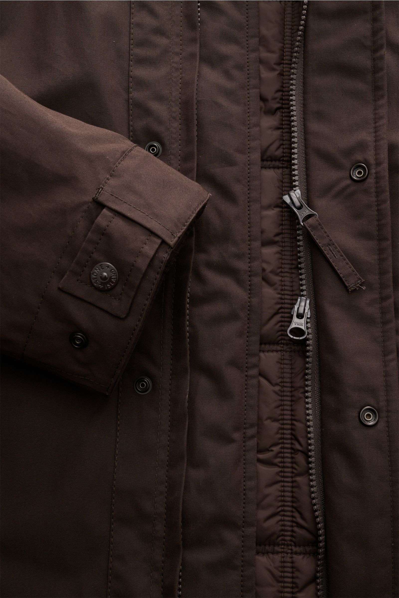 Nahaufnahme der dunkelbraunen Stone Island Jacke 'Weatherproof Cotton Canvas Ghost Piece' mit Detailansicht von Reißverschluss, Knöpfen und Ärmelabschluss aus dicht gewebtem Baumwoll-Canvas.
