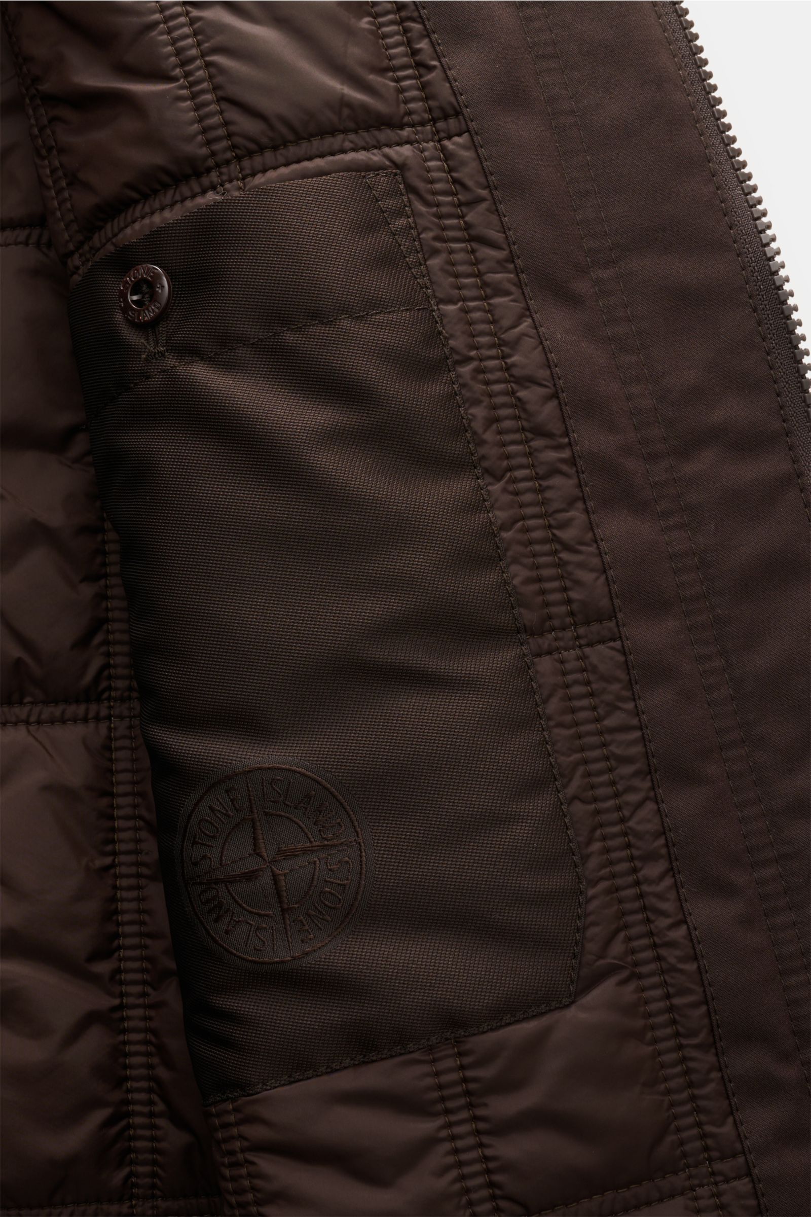 Nahaufnahme der Innentasche der Stone Island Jacke 'Weatherproof Cotton Canvas Ghost Piece' dunkelbraun, mit gestepptem Futter und typischem Kompass-Badge aus wind- und wasserabweisendem Baumwoll-Canvas.
