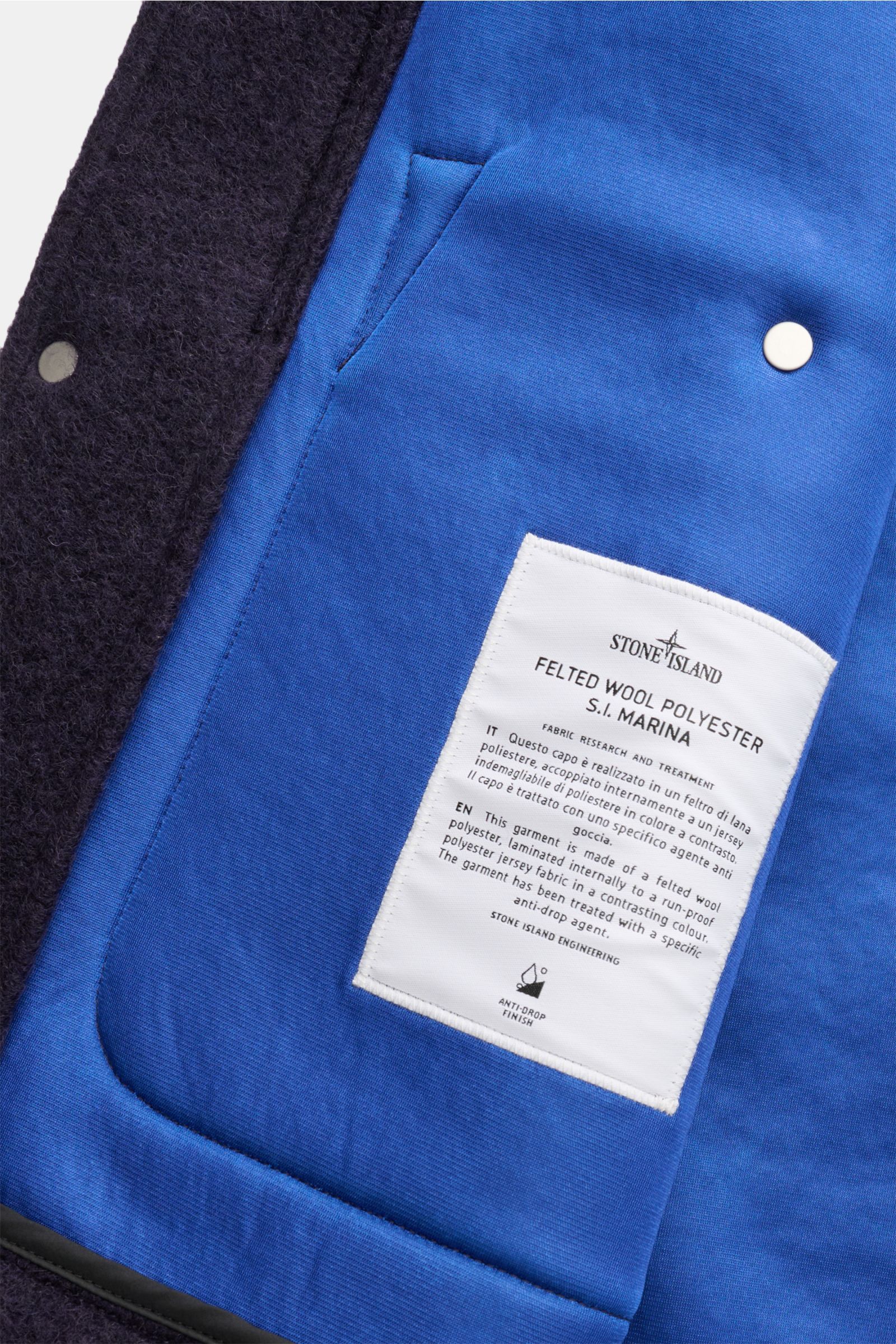 Close-up image of the inside of the Stone Island Jacke 'Marina Felted Wool Polyester S.I. Marina' navy, showing the blue inner lining with a white fabric label and part of the dark felted wool outer layer, photographed from a top-down angle. -
